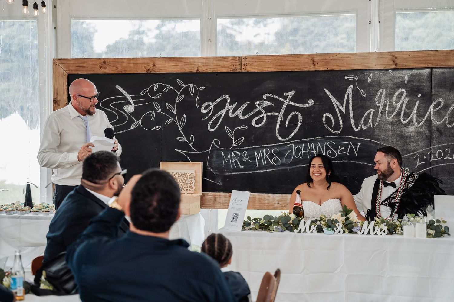 Best man gives speech at wedding reception before couple seated at table with Just Married chalkboard.
