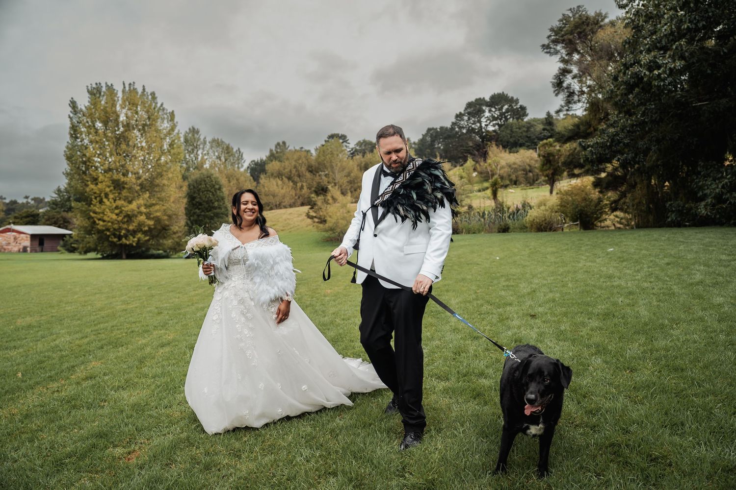 Newlywed couple walking black dog across green lawn on cloudy wedding day outdoors.
