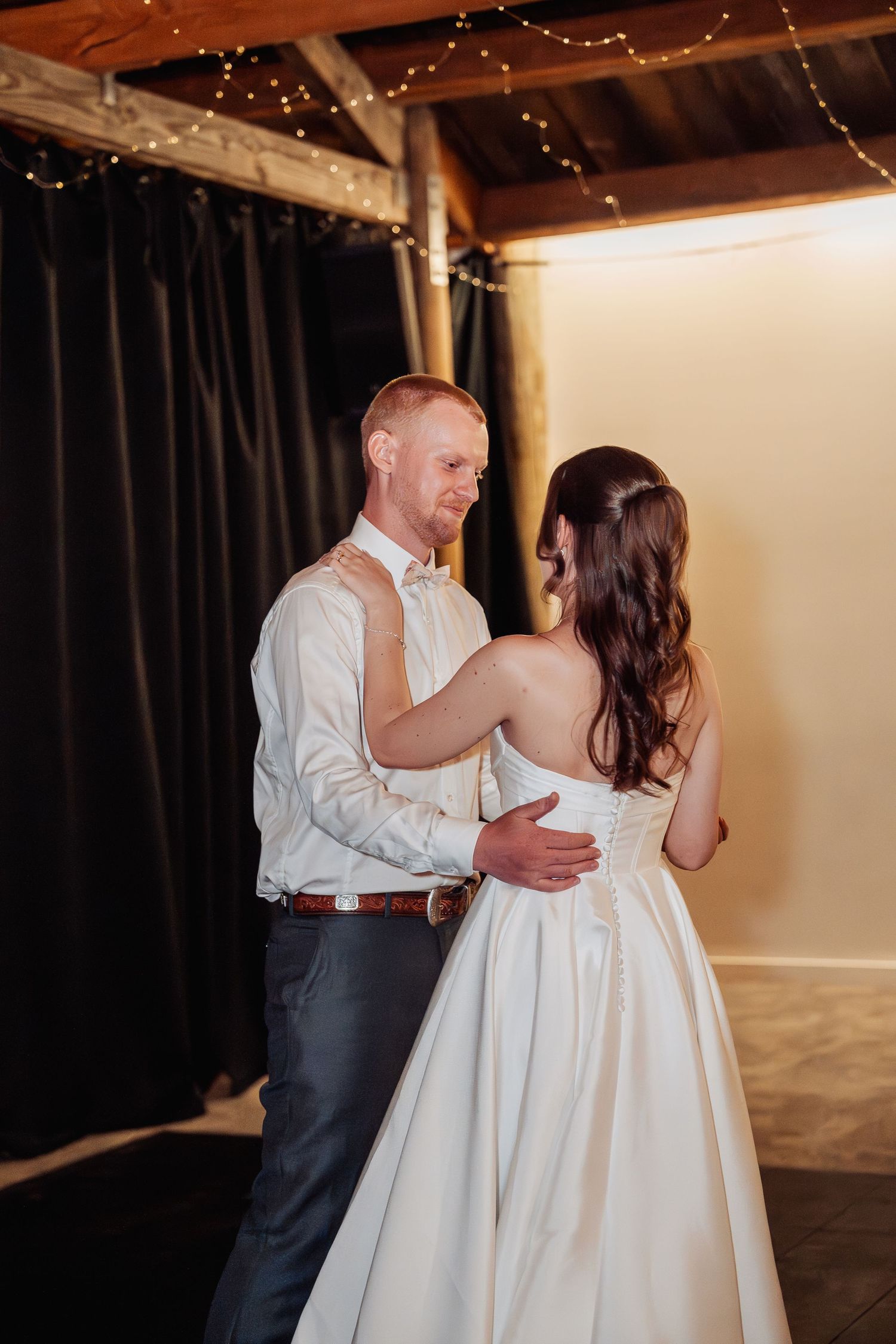Bride and groom share their first dance under warm fairy lights in a rustic barn.