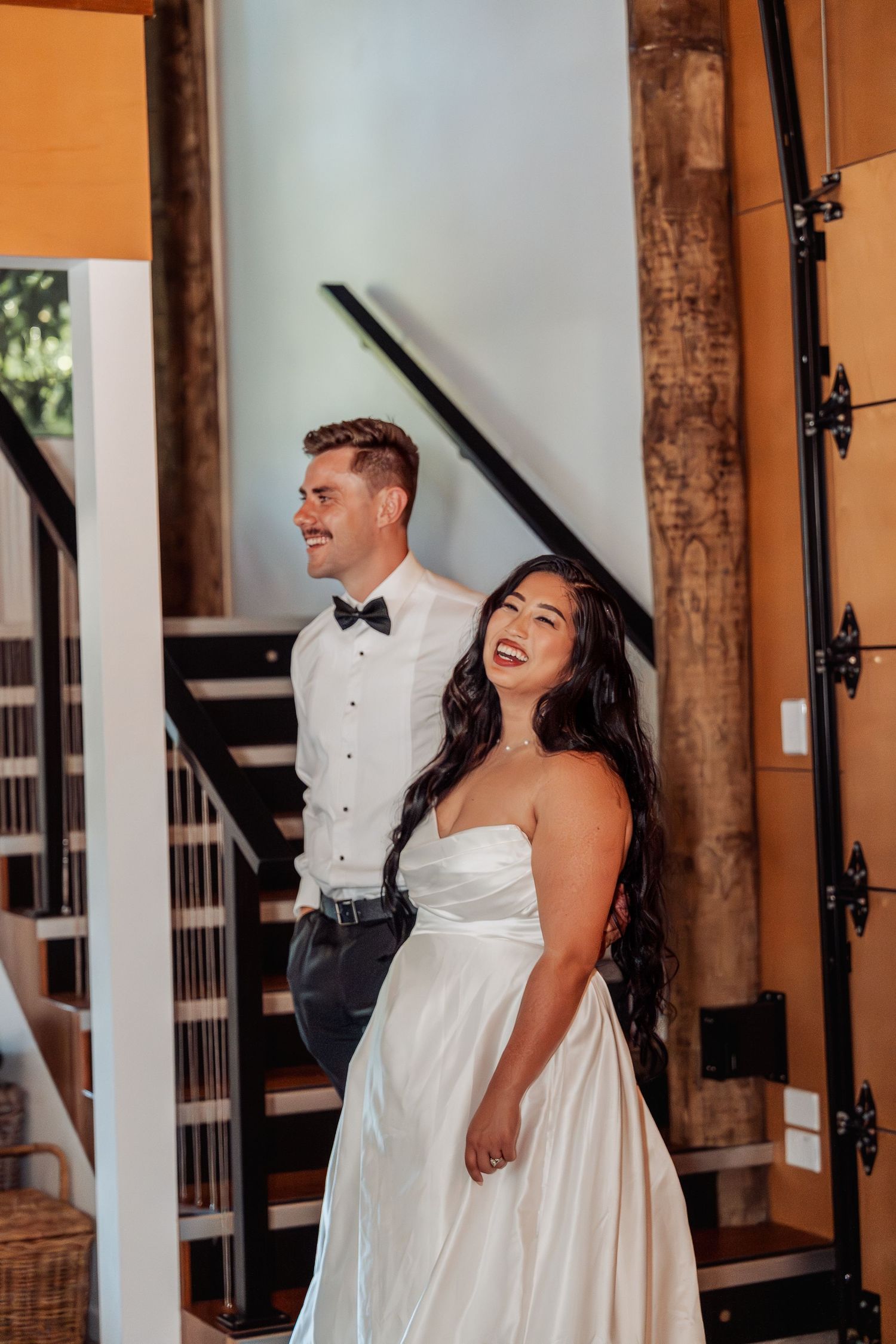 Smiling bride in strapless white satin gown stands with groom in bow tie inside a rustic venue with wooden staircase.