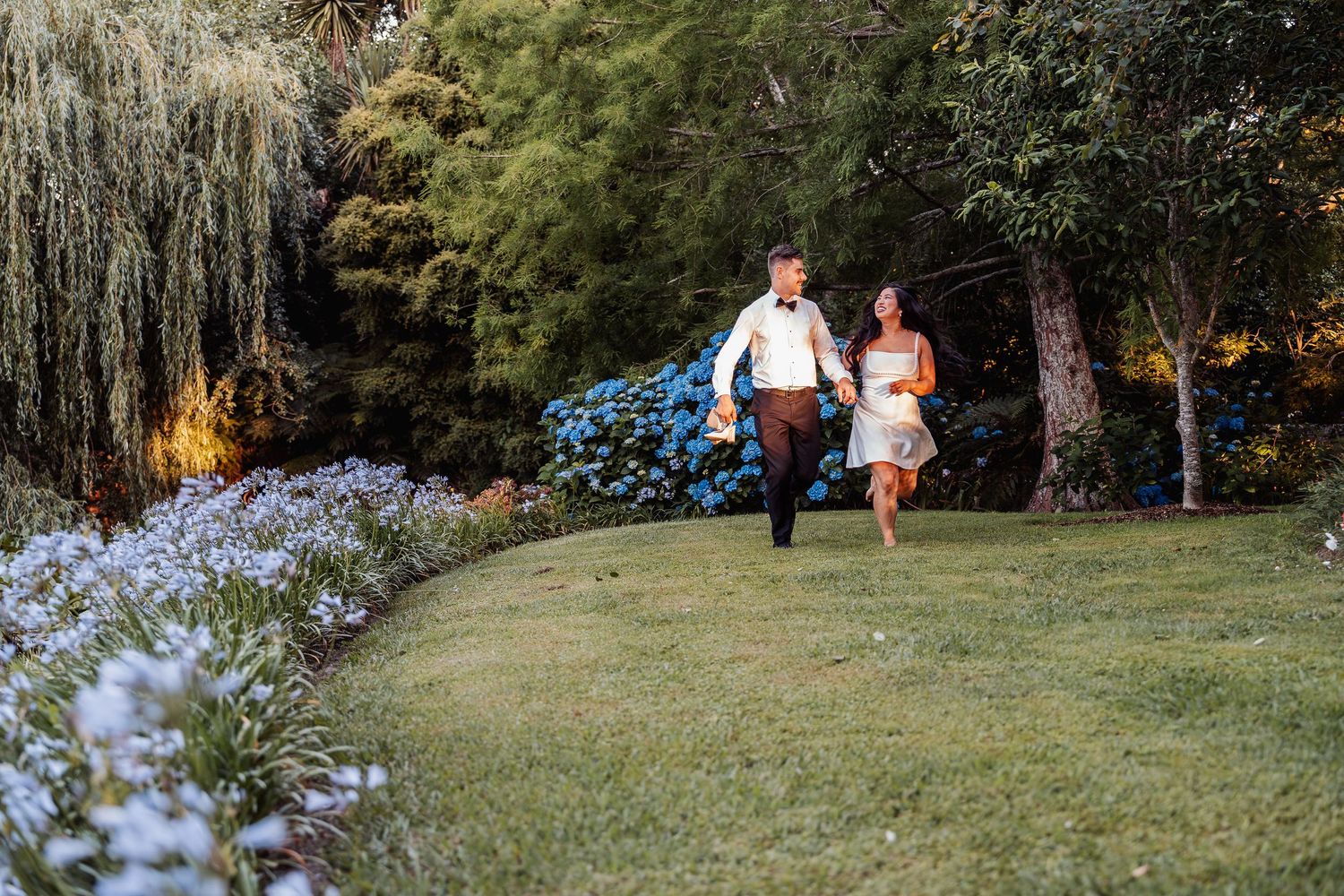 Bride and groom holding hands walking through a lush garden with colorful flowers at dusk.
