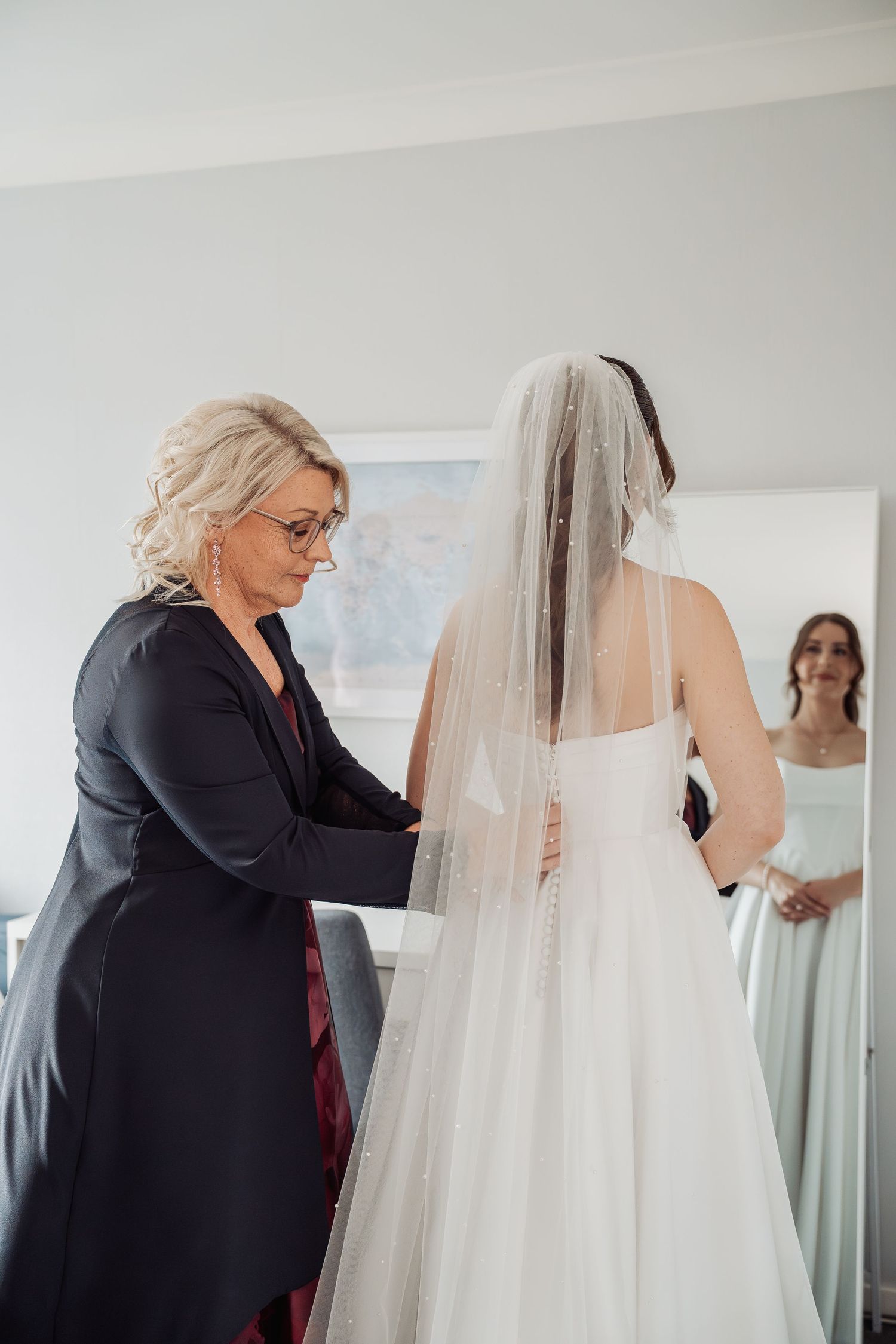 Mother helping bride with wedding dress zipper while bridesmaid watches in mirror reflection.