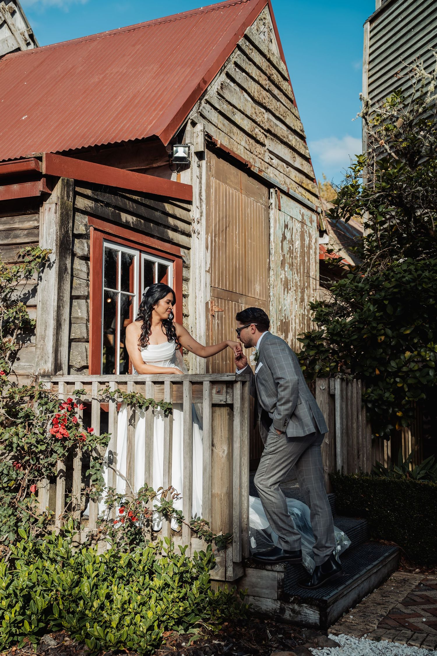 Groom kisses bride's hand over white picket fence beside rustic wooden cottage.