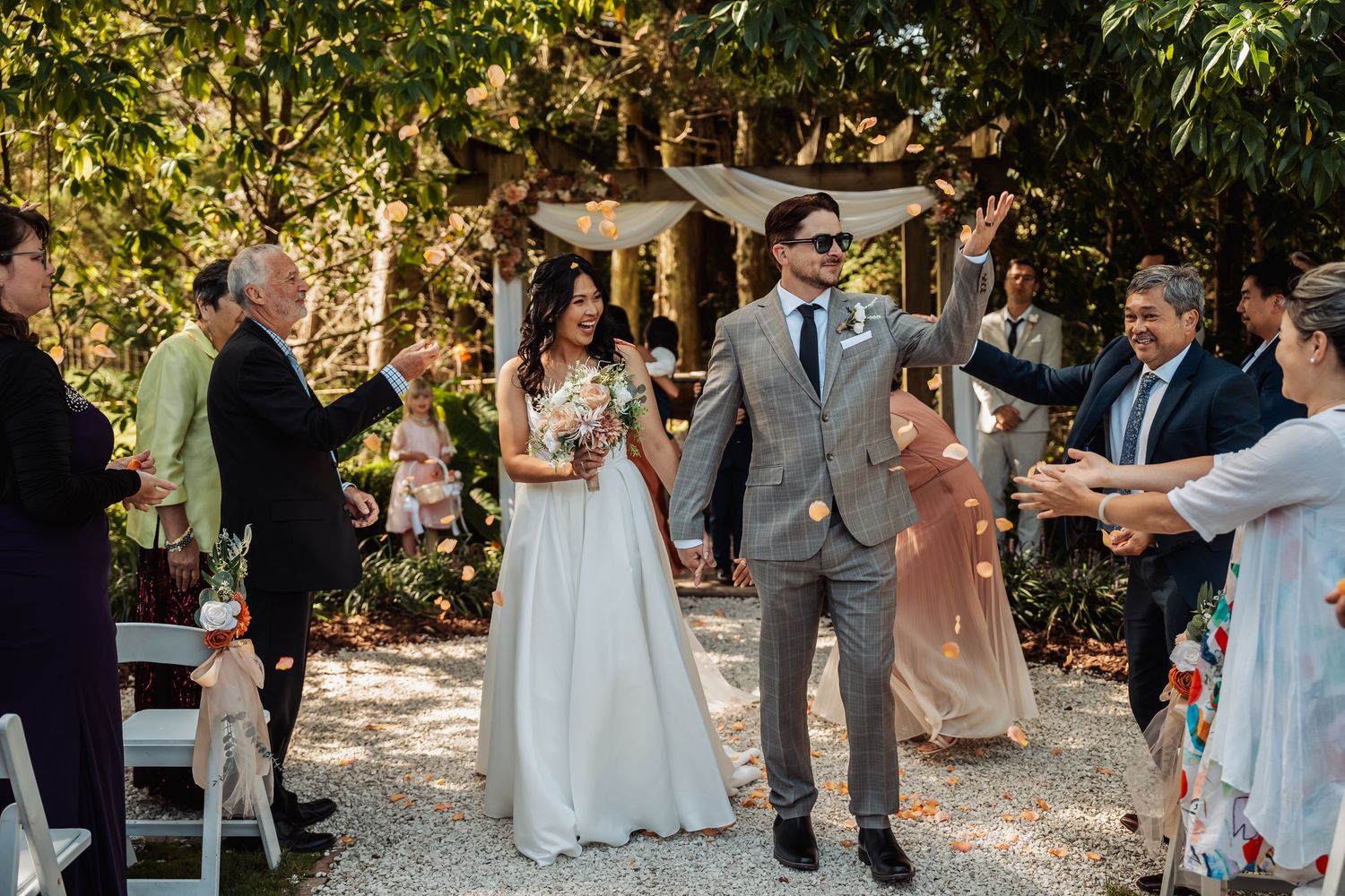Bride and groom walk down outdoor wedding aisle as guests toss flower petals in a garden ceremony.