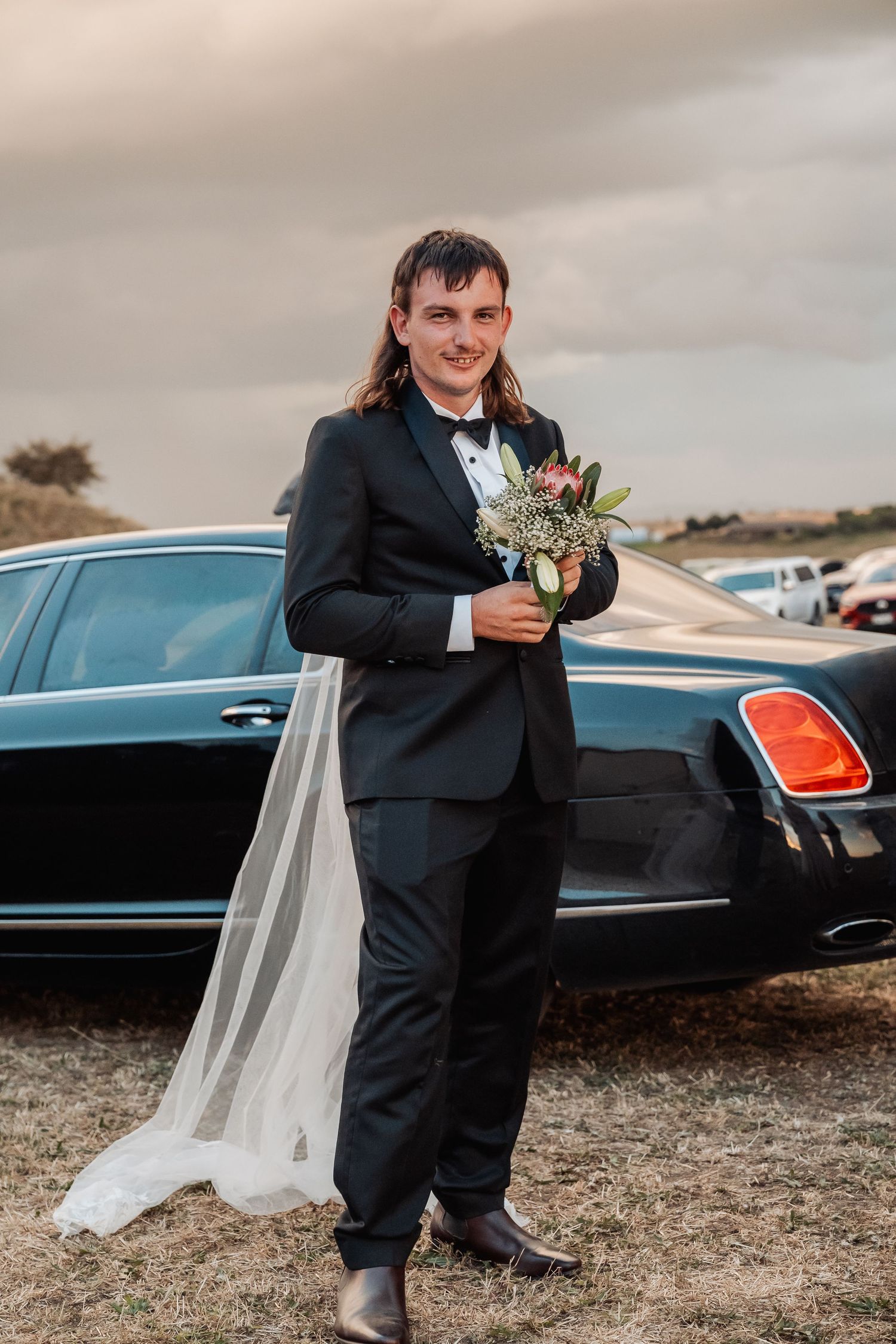 Groom in black tuxedo holding a bridal bouquet and veil while standing in front of a black luxury car.