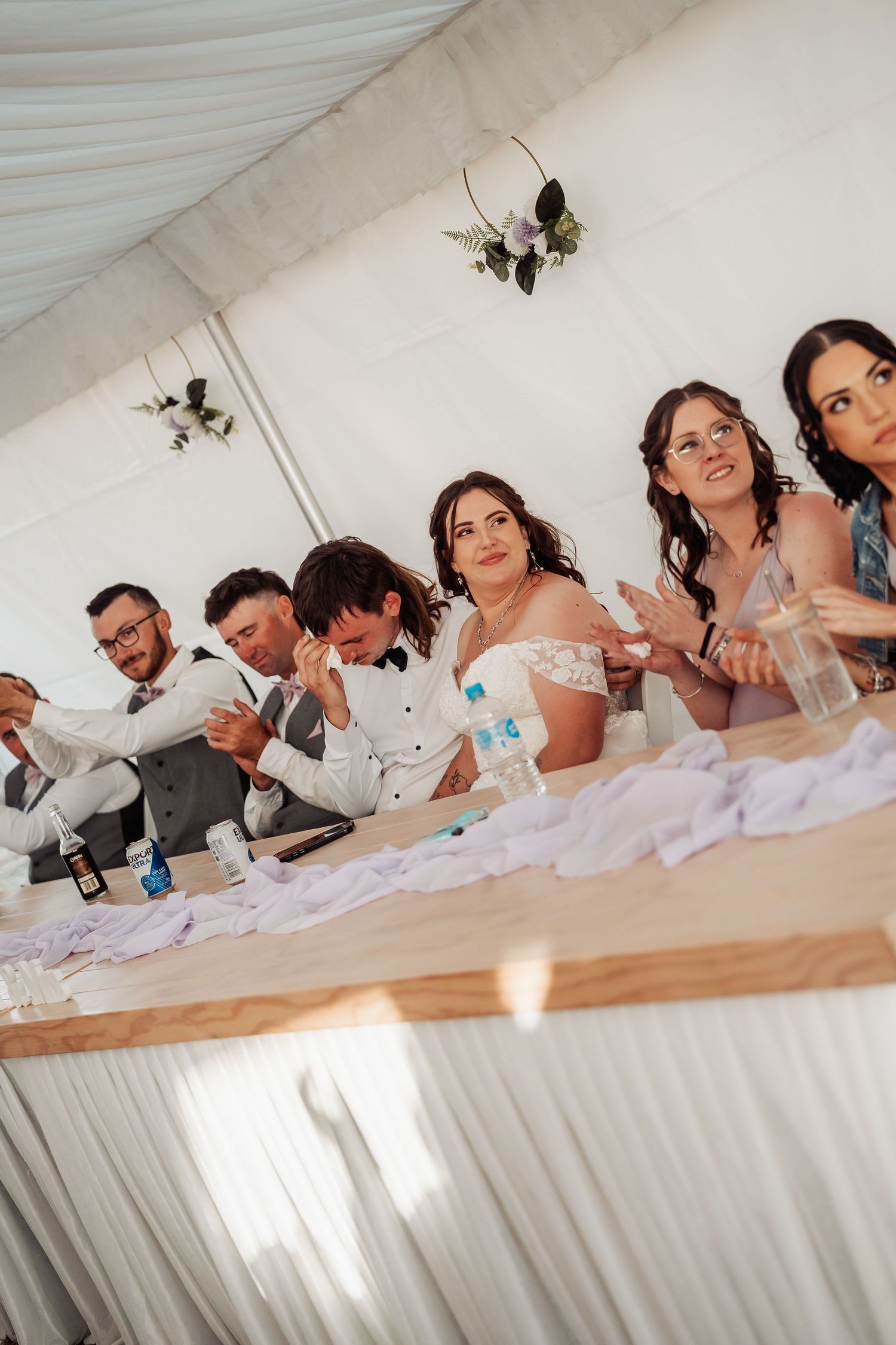 Wedding party sits at head table under white tent, bride and groom share an emotional moment.