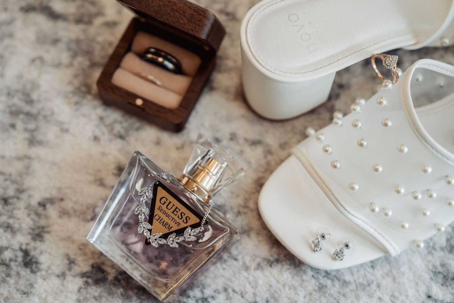 Guess perfume bottle with wedding rings in wooden box and pearl-studded white bridal sandals on marble surface.