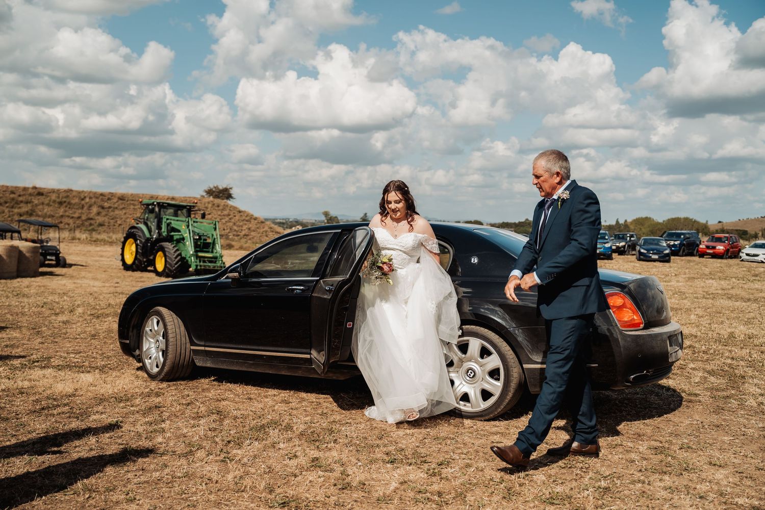 Bride in white gown arriving at outdoor wedding venue in black luxury car with escort.