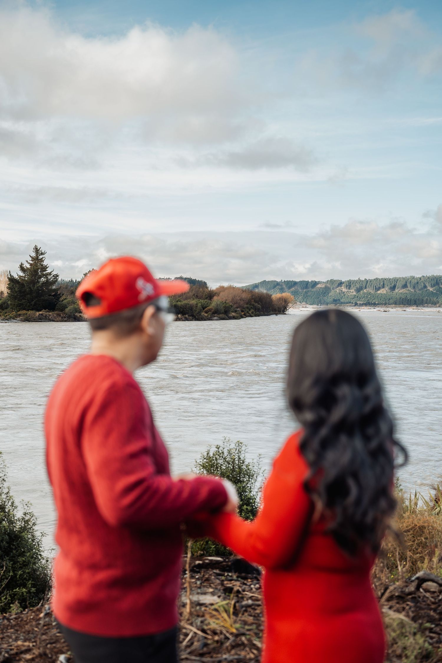 Couple in matching red outfits holding hands while gazing at a scenic river landscape under a cloudy sky.
