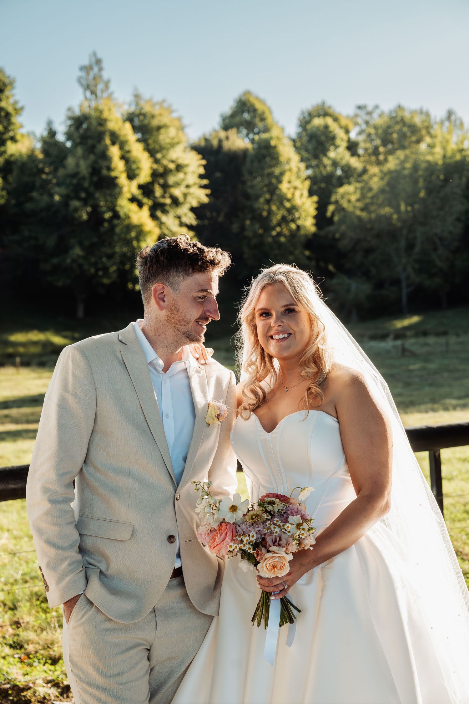 Smiling bride in strapless gown holds bouquet beside groom in beige suit at outdoor wedding.