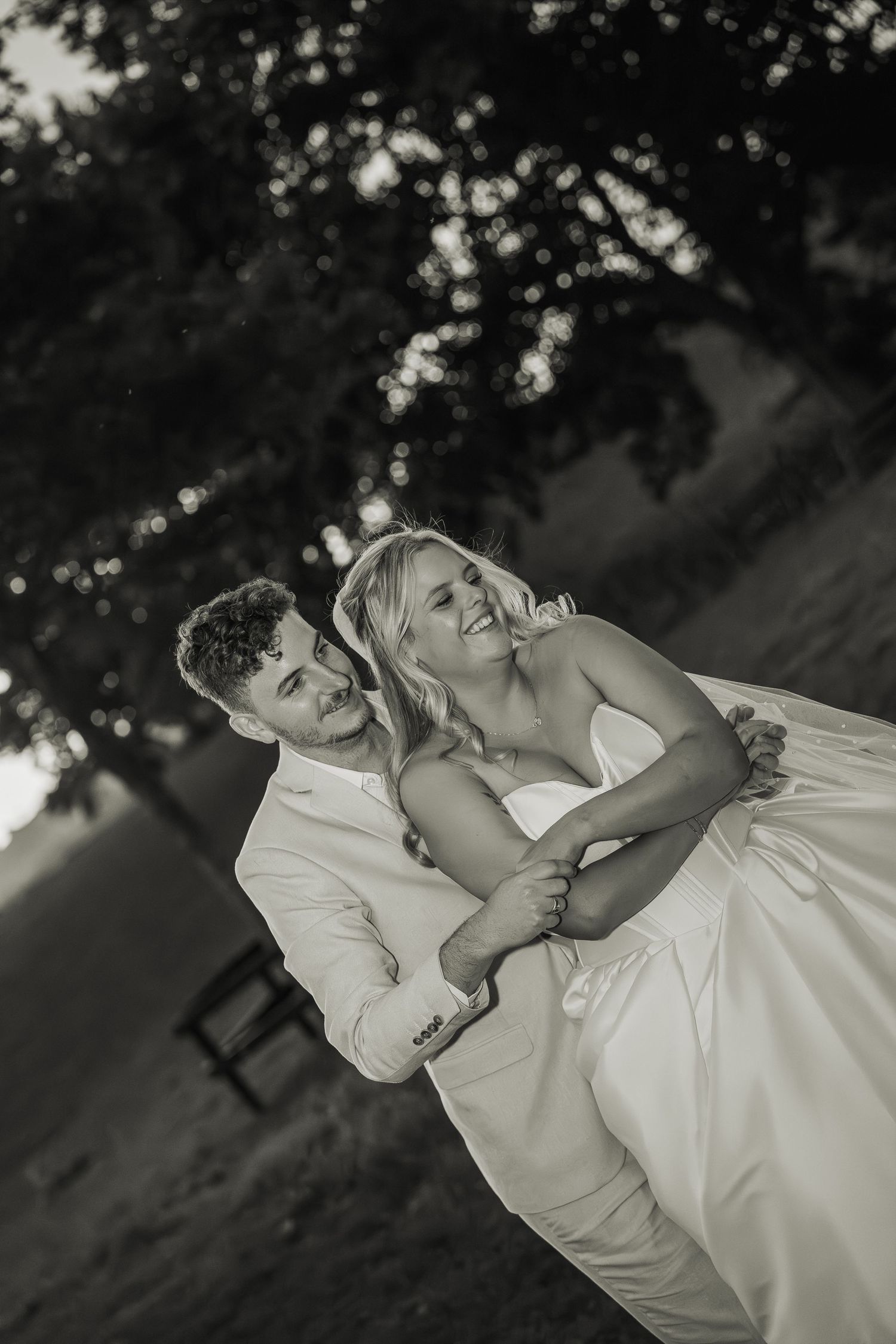 Black and white photo of groom lifting laughing bride outdoors under a shady tree.