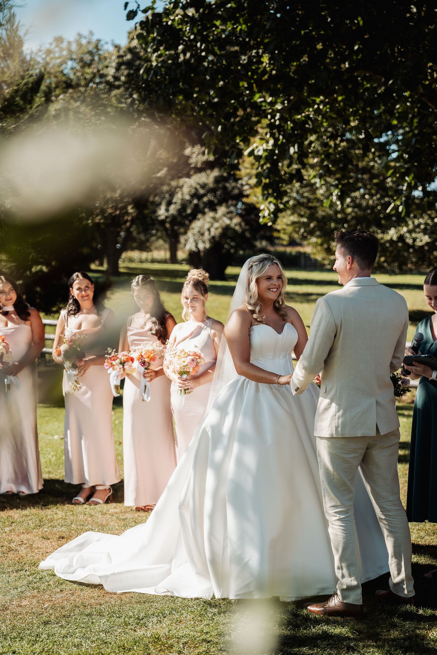 Bride in white ball gown smiling at groom during outdoor wedding ceremony with bridesmaids in blush pink.