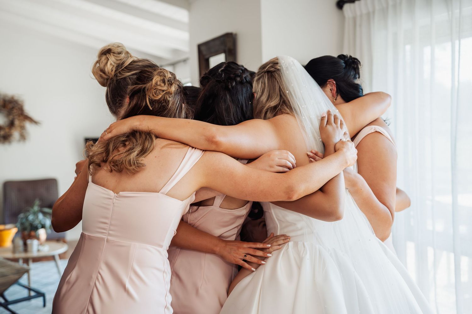 Bride in white wedding dress sharing a group hug with bridesmaids in blush pink outfits indoors.