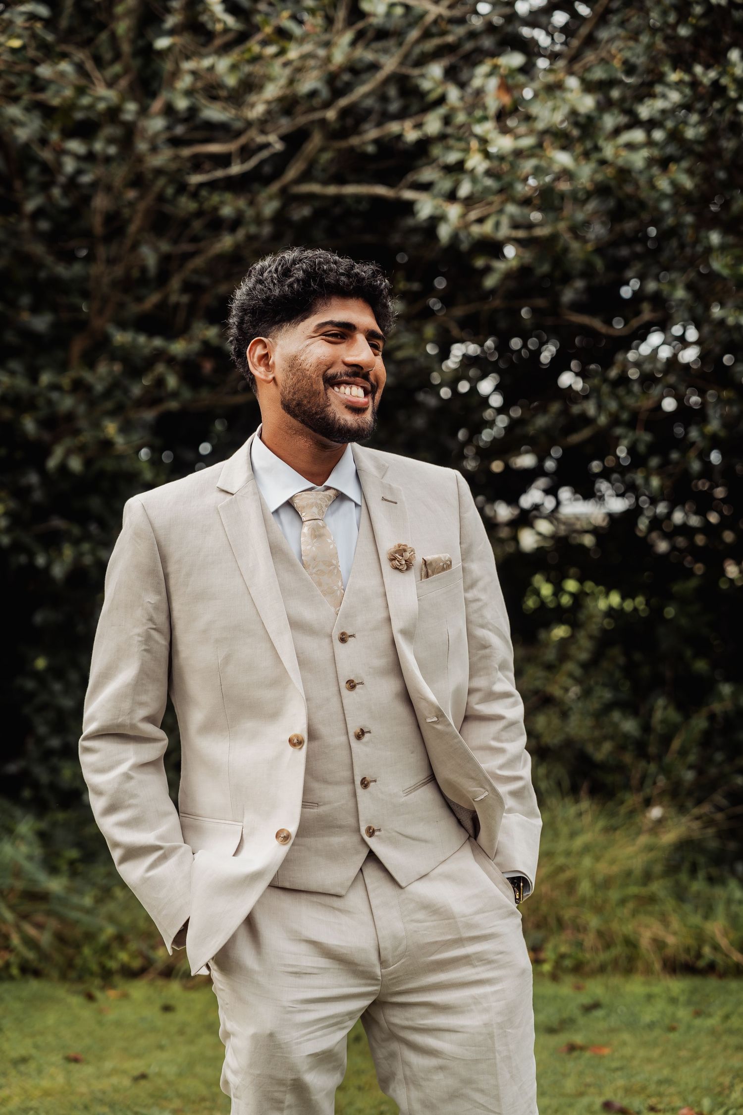 Handsome groom laughing in elegant cream three-piece suit with tie and boutonniere outdoors.
