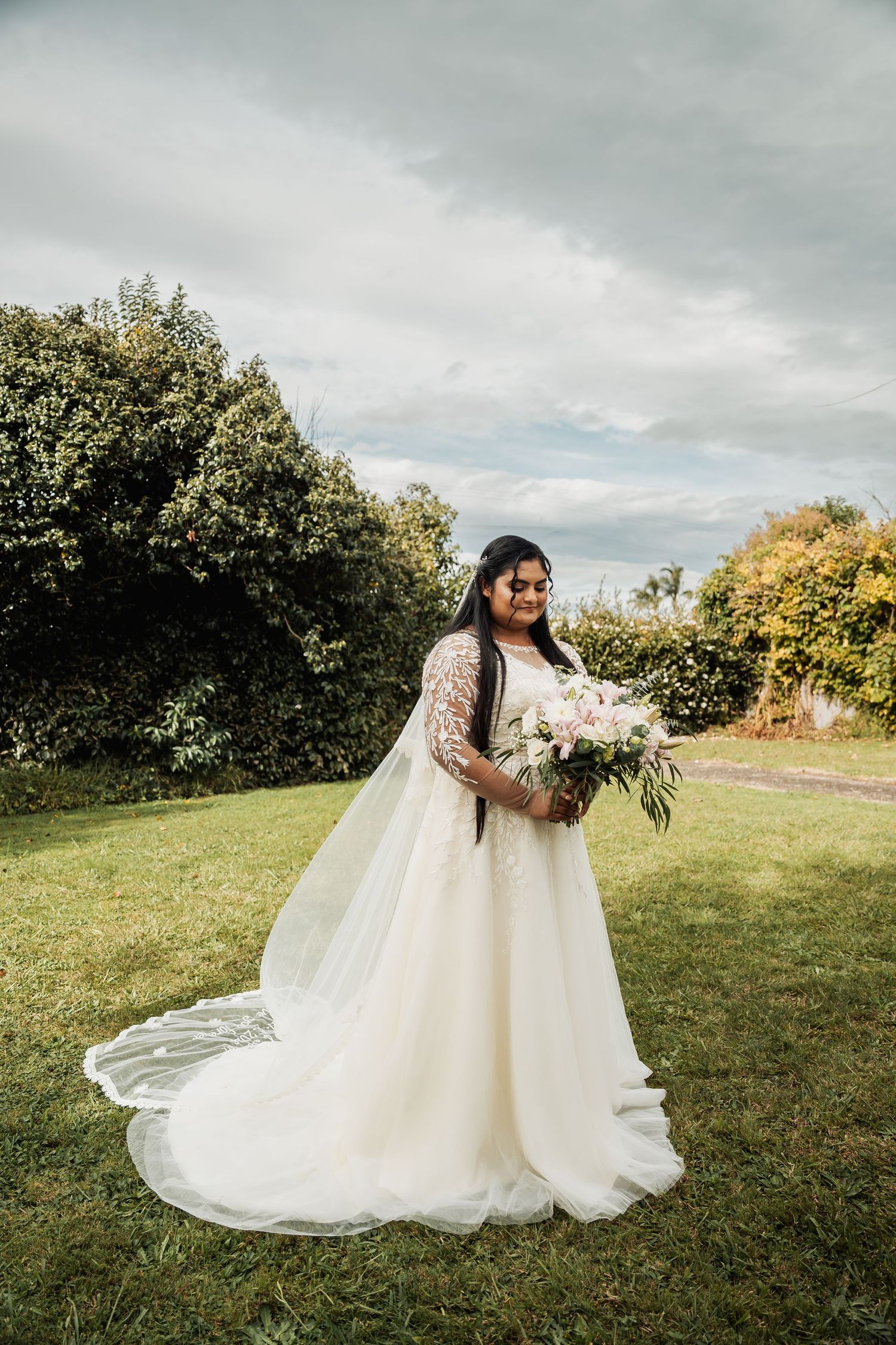 Bride in a flowing white lace wedding gown holding a pink bouquet, standing on green grass with trees behind her.