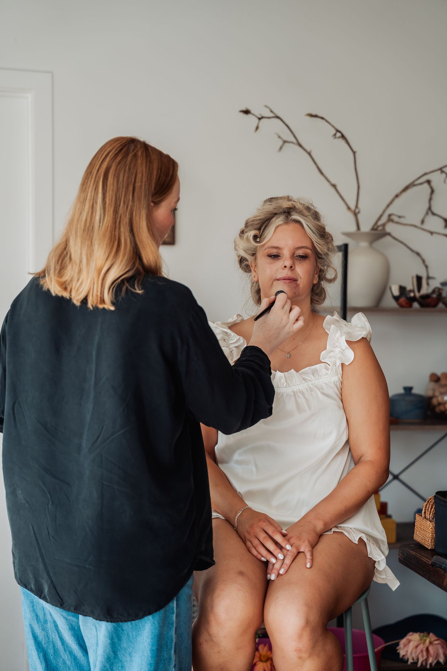 Makeup artist applying lipstick to a blonde bride in hair rollers wearing a white dress before her wedding.