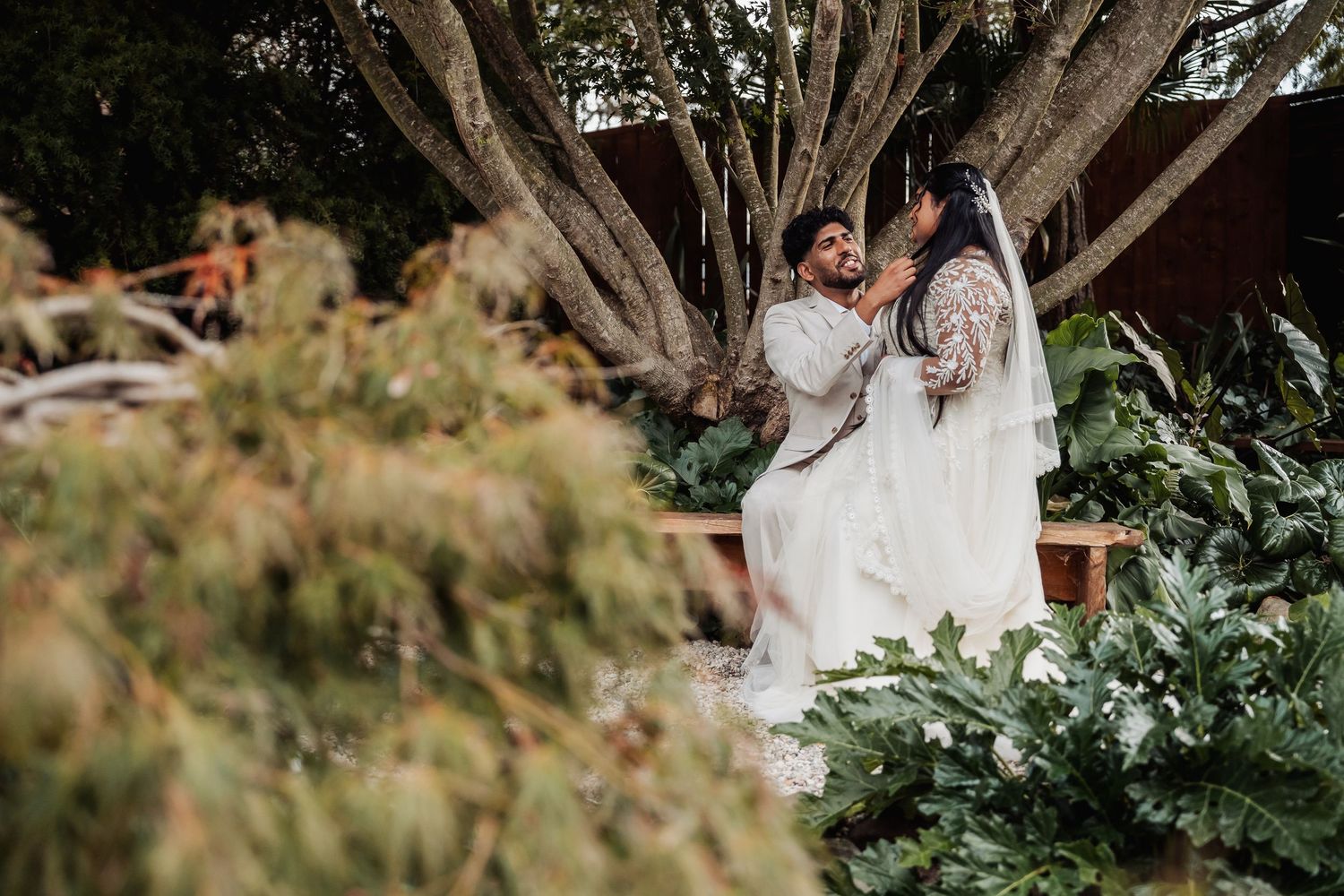 Bride and groom sharing intimate moment seated among lush tropical greenery and large tree.