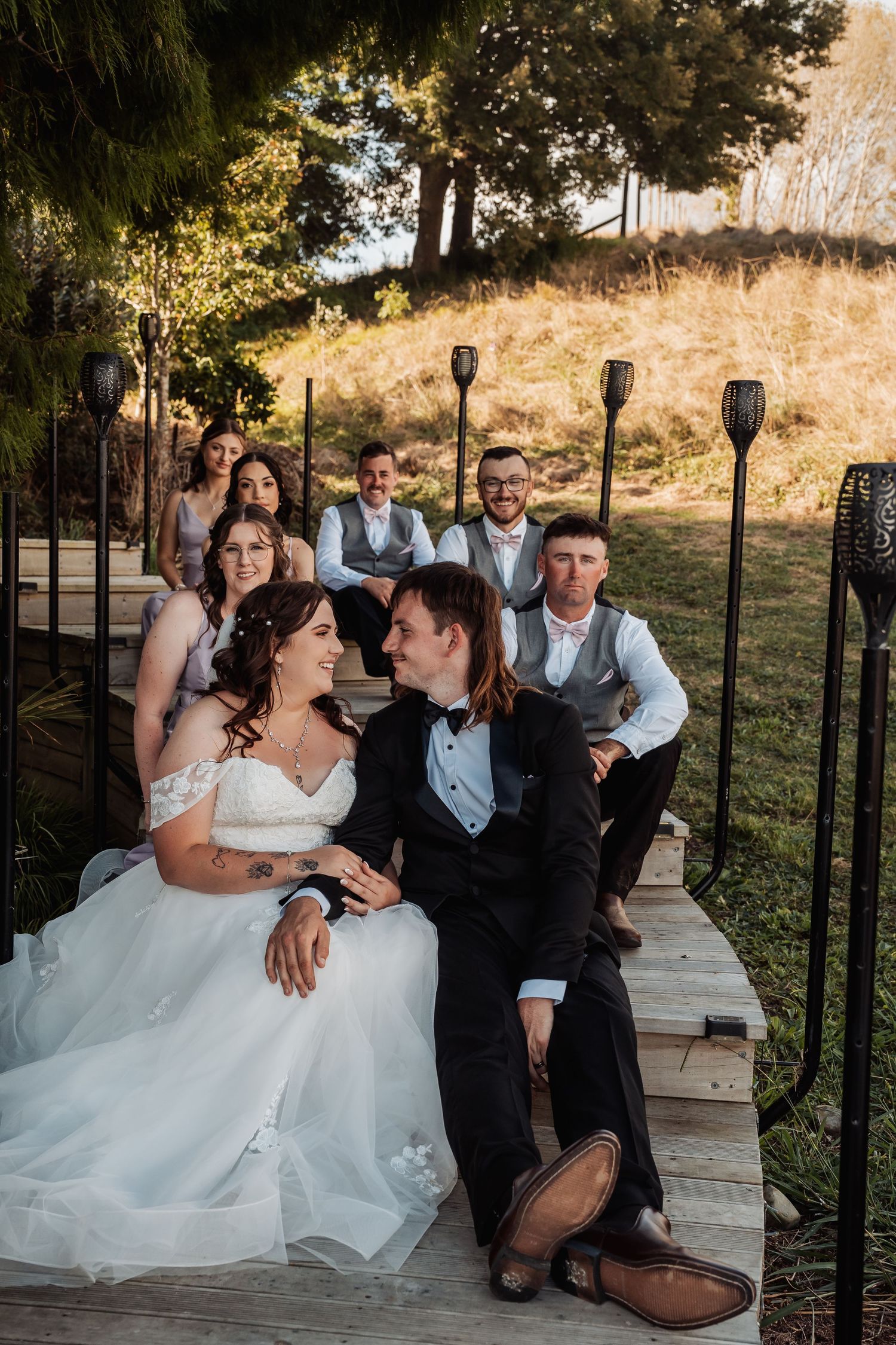 Wedding party relaxing on wooden steps outdoors, bride and groom gazing at each other, bridesmaids and groomsmen seated behind.