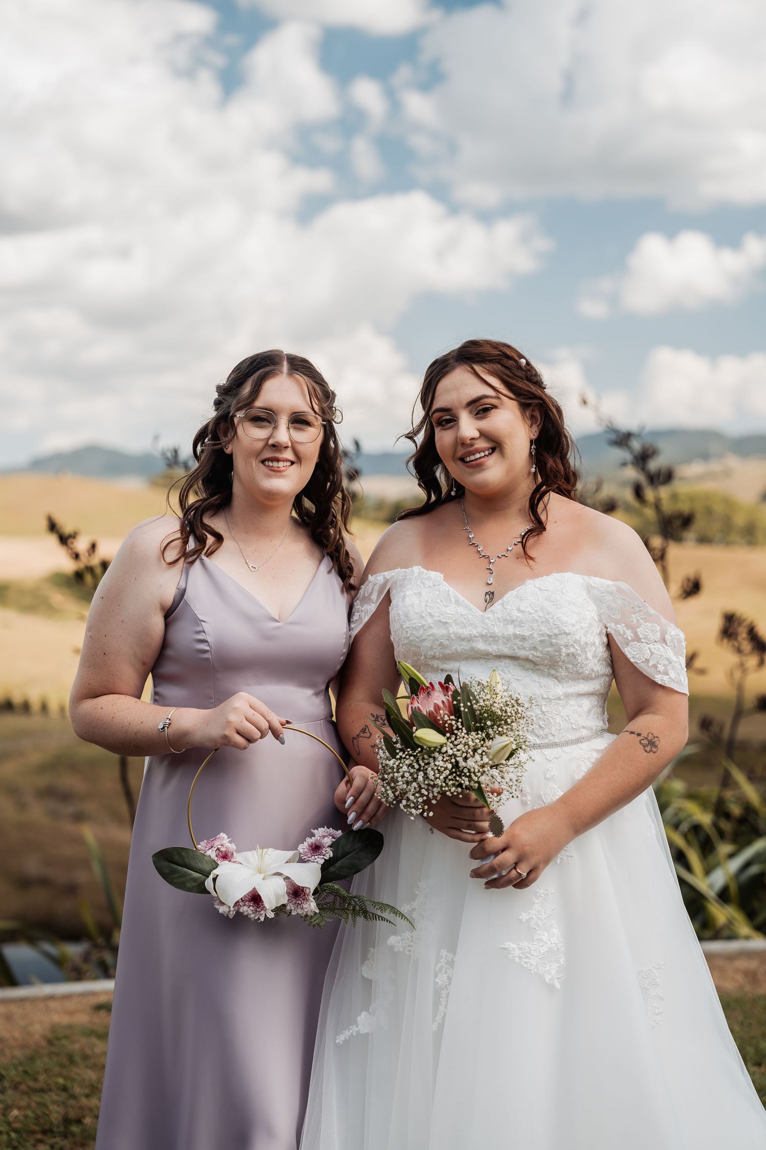 Bride in lace off-shoulder gown poses with bridesmaid in lavender dress holding bouquets.
