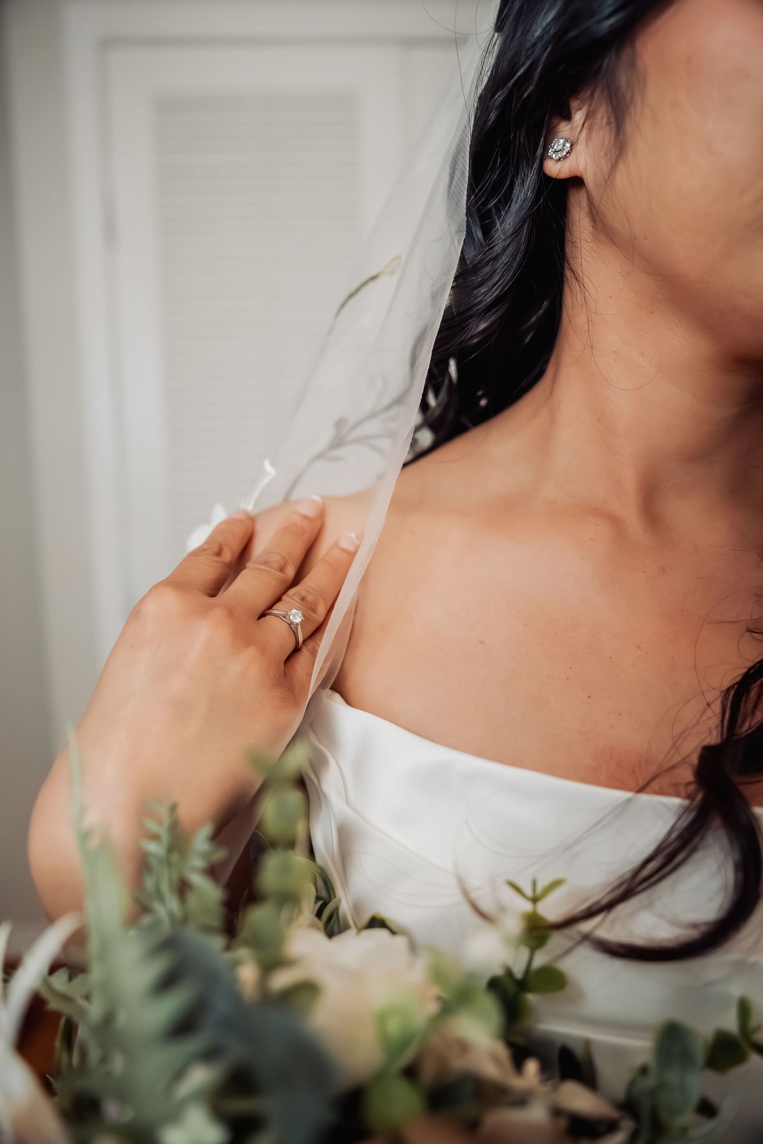 Close-up of bride in white strapless gown holding veil, wearing diamond ring and stud earrings with bridal bouquet.