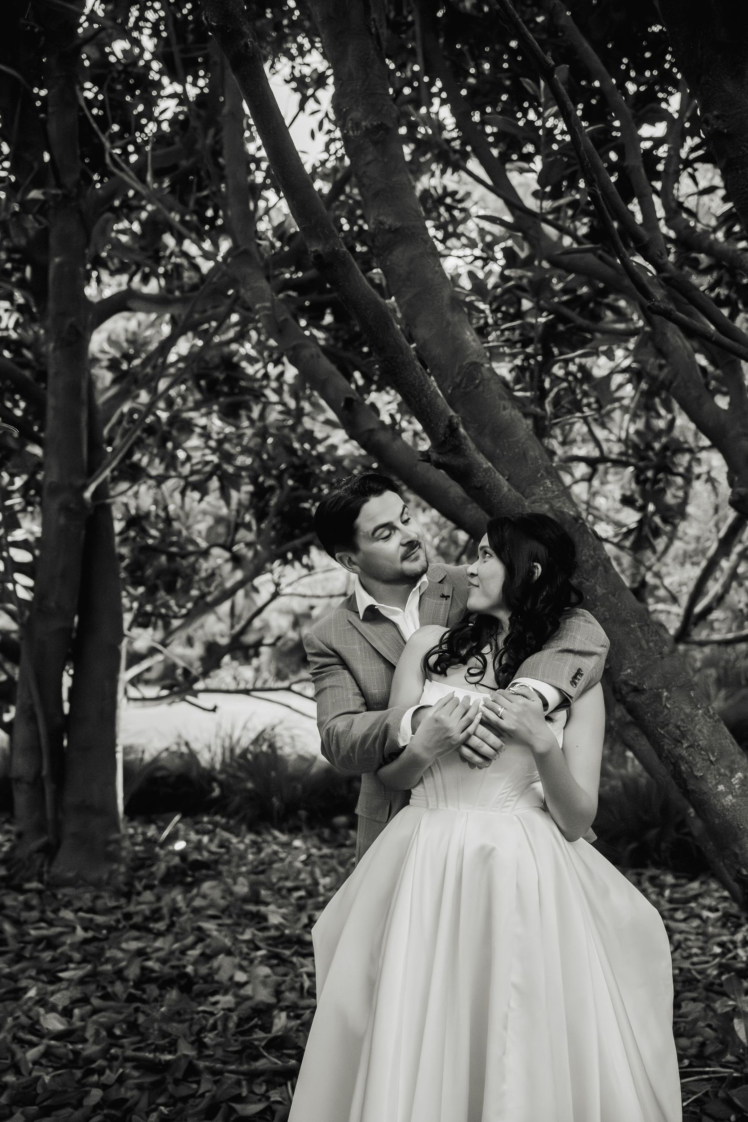 Black and white romantic portrait of bride and groom embracing under trees in a lush garden wedding setting.