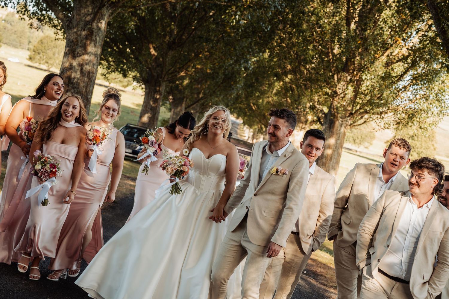 Wedding party in beige and blush attire walking together under shaded trees.