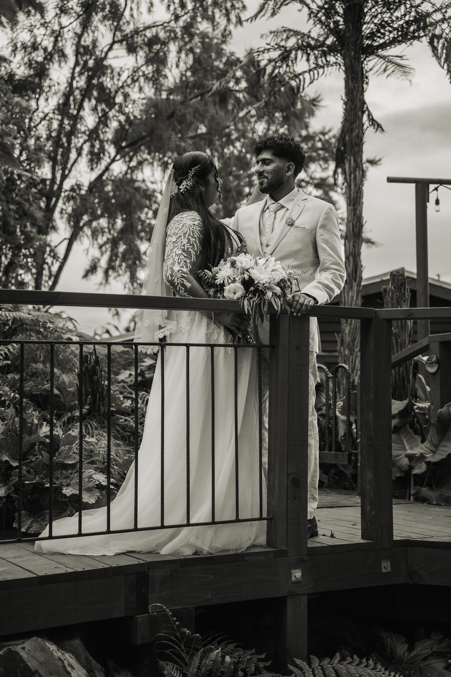 Black and white photo of bride and groom on wooden bridge surrounded by trees.
