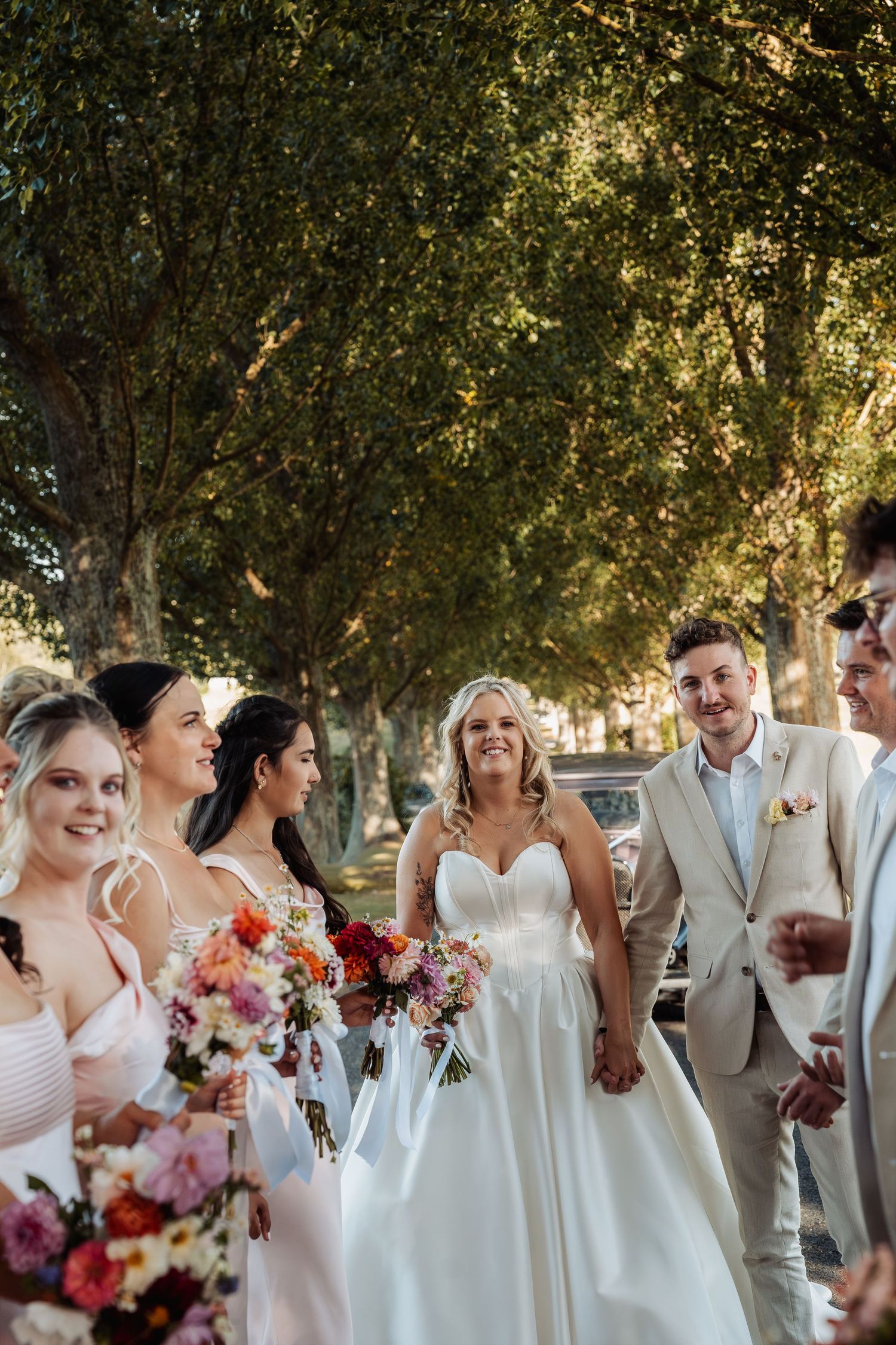 Bride holding hands with groom surrounded by smiling bridesmaids with colorful bouquets outdoors.