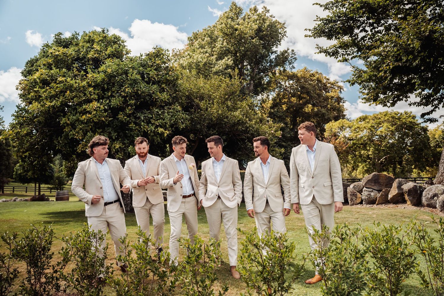 Six groomsmen in matching beige suits walking together outdoors on a sunny day at a wedding venue.