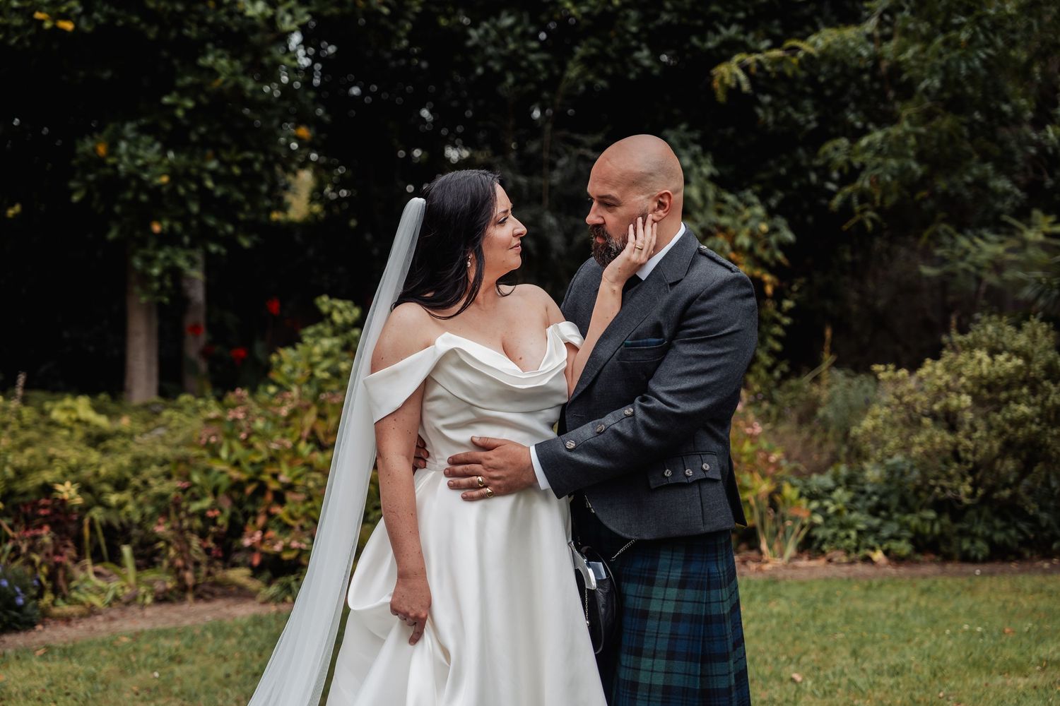Bride and groom share a tender moment outdoors, she in an off-shoulder gown with veil, he in a tartan kilt.