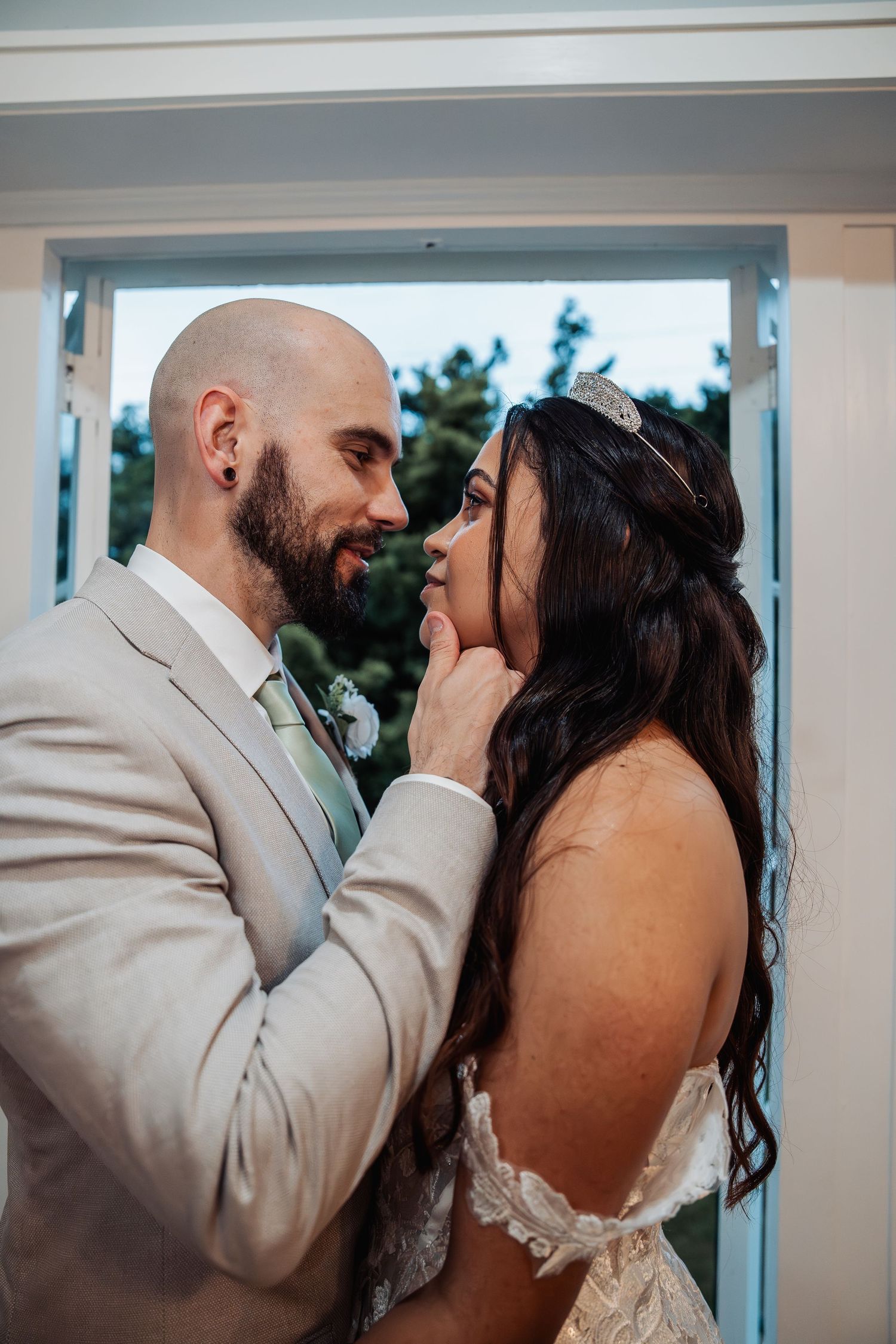 A groom in a beige suit tenderly touches his bride's face as they share an intimate moment near a window.