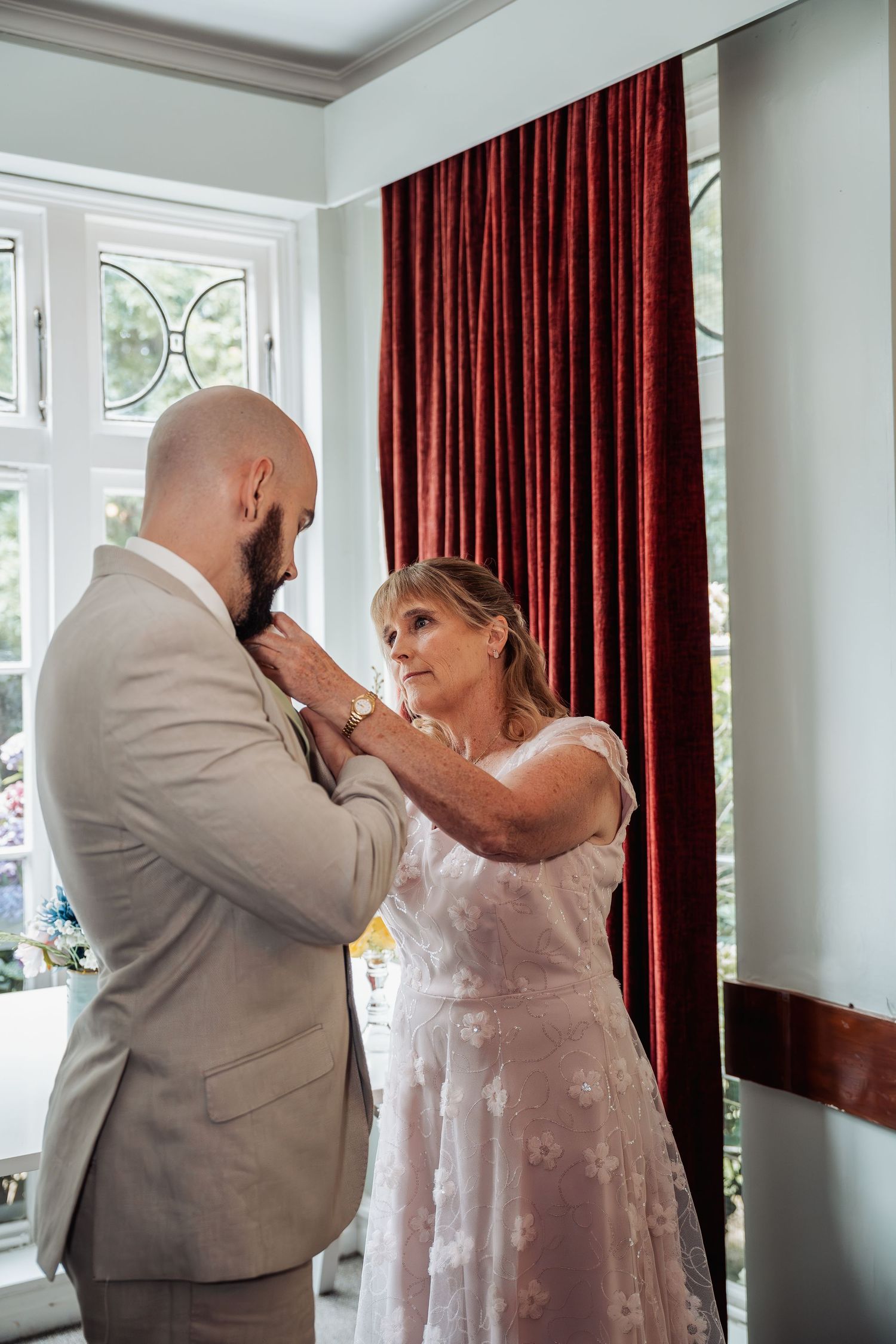 Woman helping bald groom adjust his tie near a red velvet curtain.