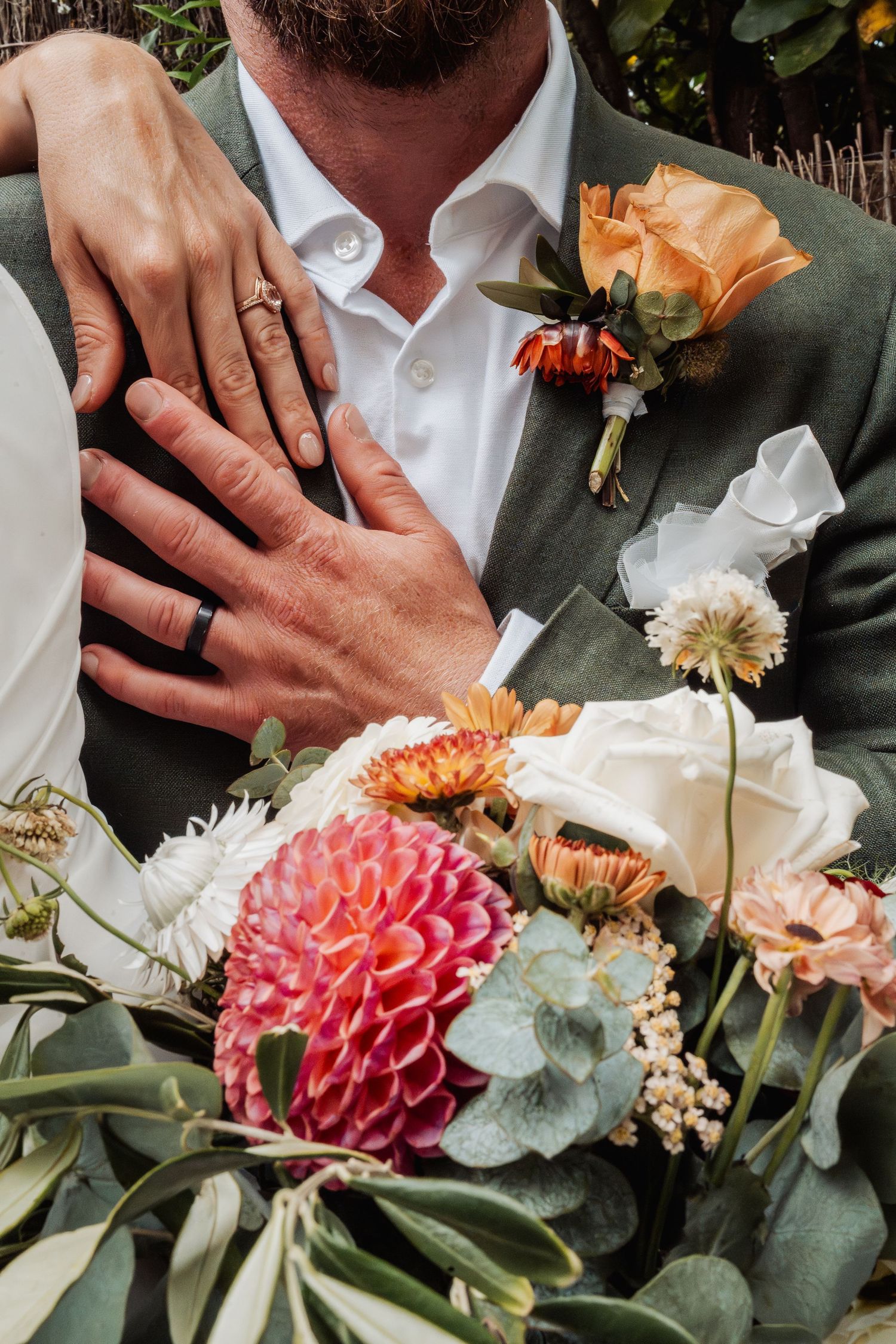 Close-up of newlyweds hands with wedding rings resting on groom's green suit beside a colorful bridal bouquet.