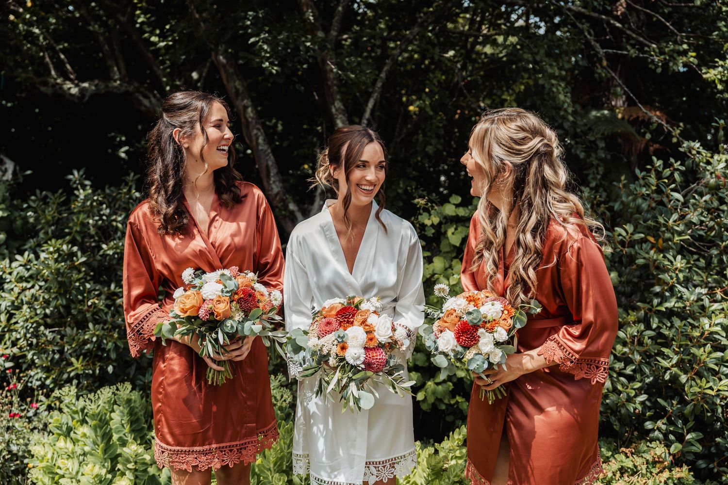 Bride in white robe flanked by two bridesmaids in rust satin robes holding colorful bouquets.