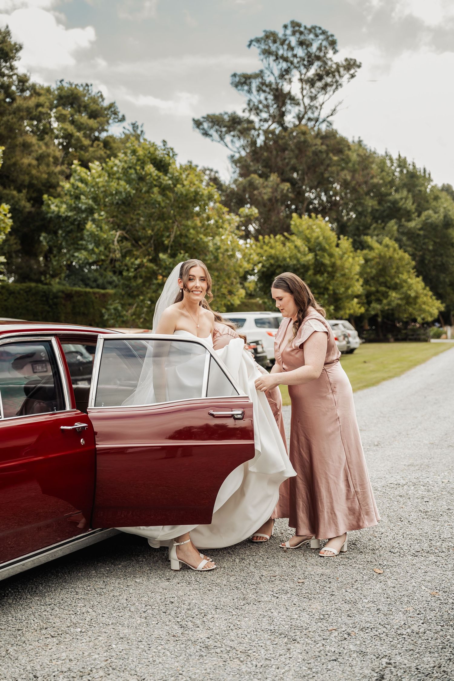 Bridesmaid in pink dress helps bride in white gown exit a classic red vintage car outdoors.