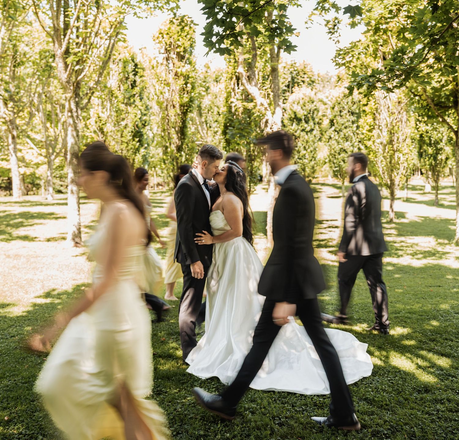 Wedding party walks through sunlit tree-lined garden as bride and groom share a romantic moment in the background.