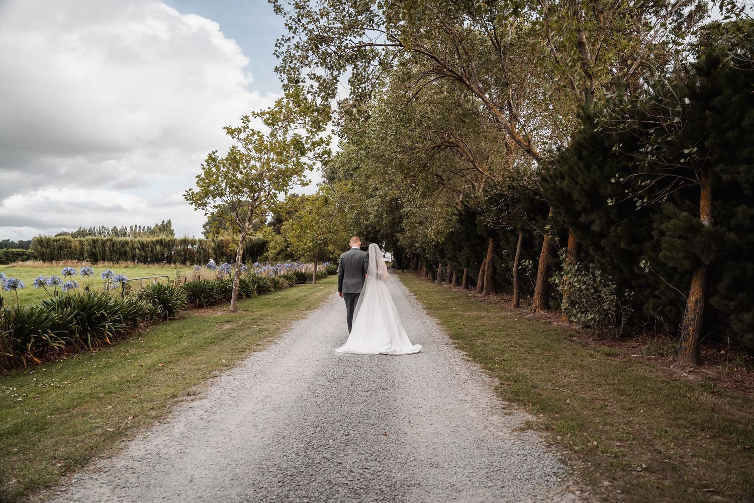 Bride and groom walk hand-in-hand down a tree-lined gravel path after their wedding.