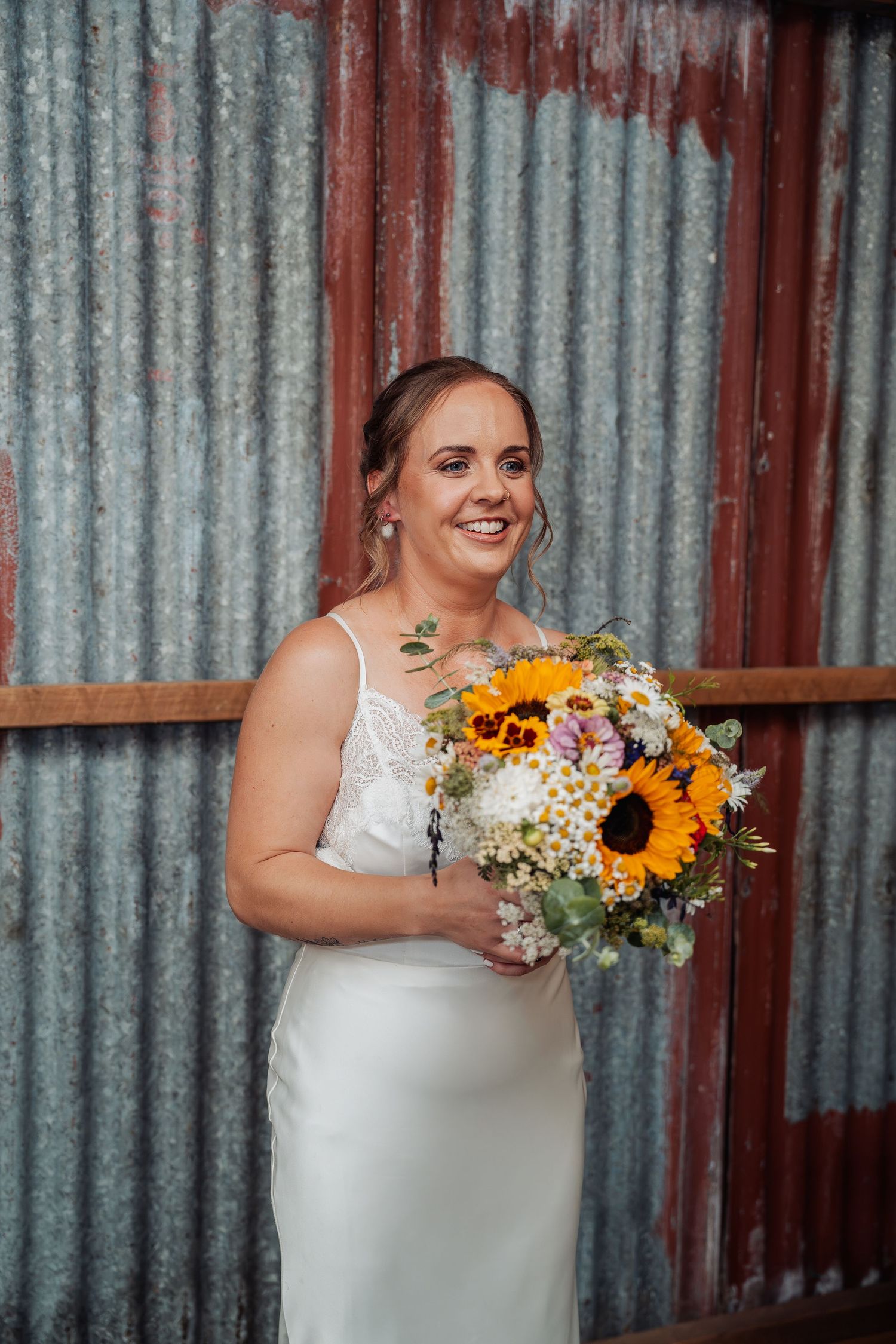 Smiling bride in a white satin dress holding a vibrant sunflower and wildflower bouquet.