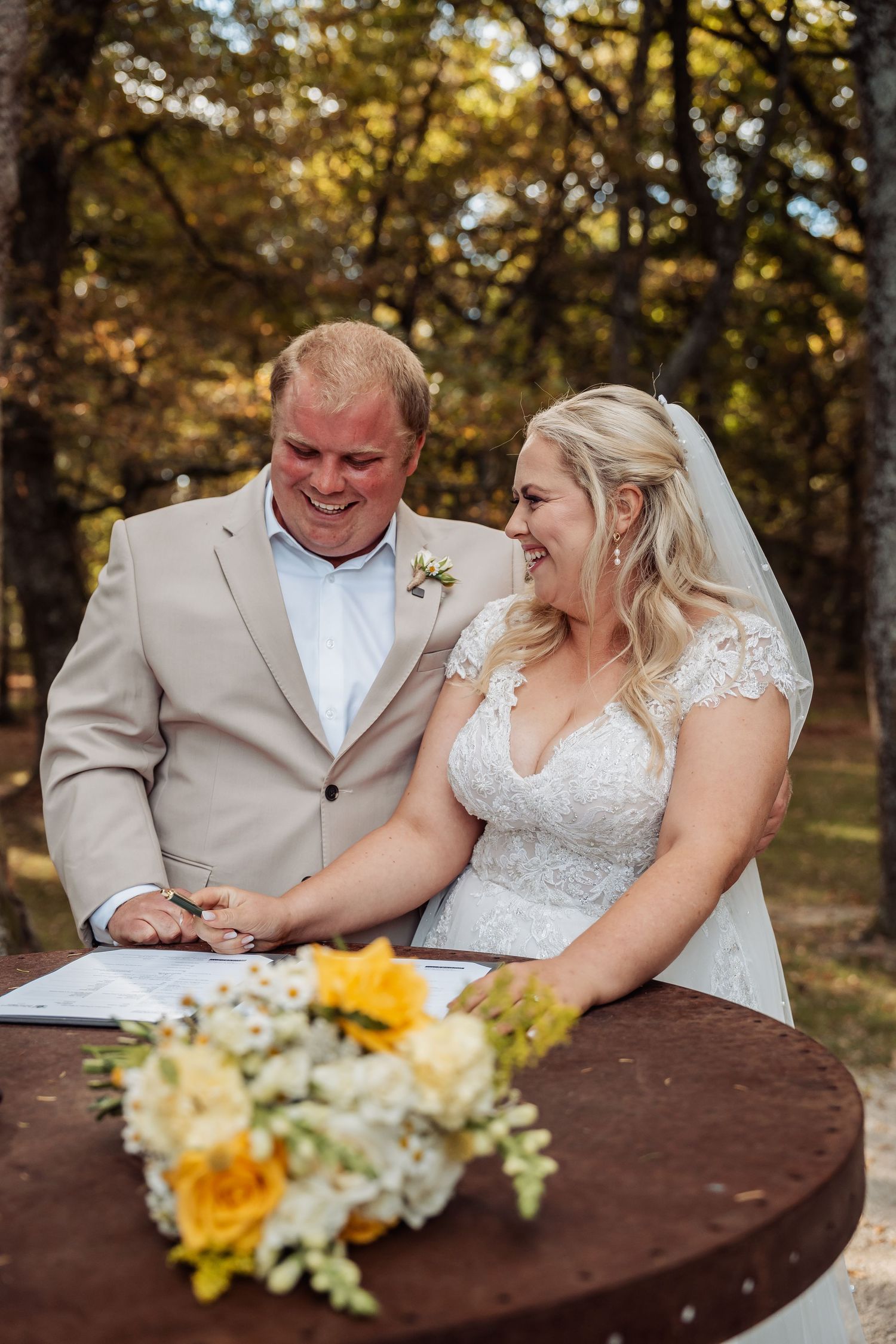 Smiling bride and groom sign wedding certificate at rustic table with yellow floral centerpiece.
