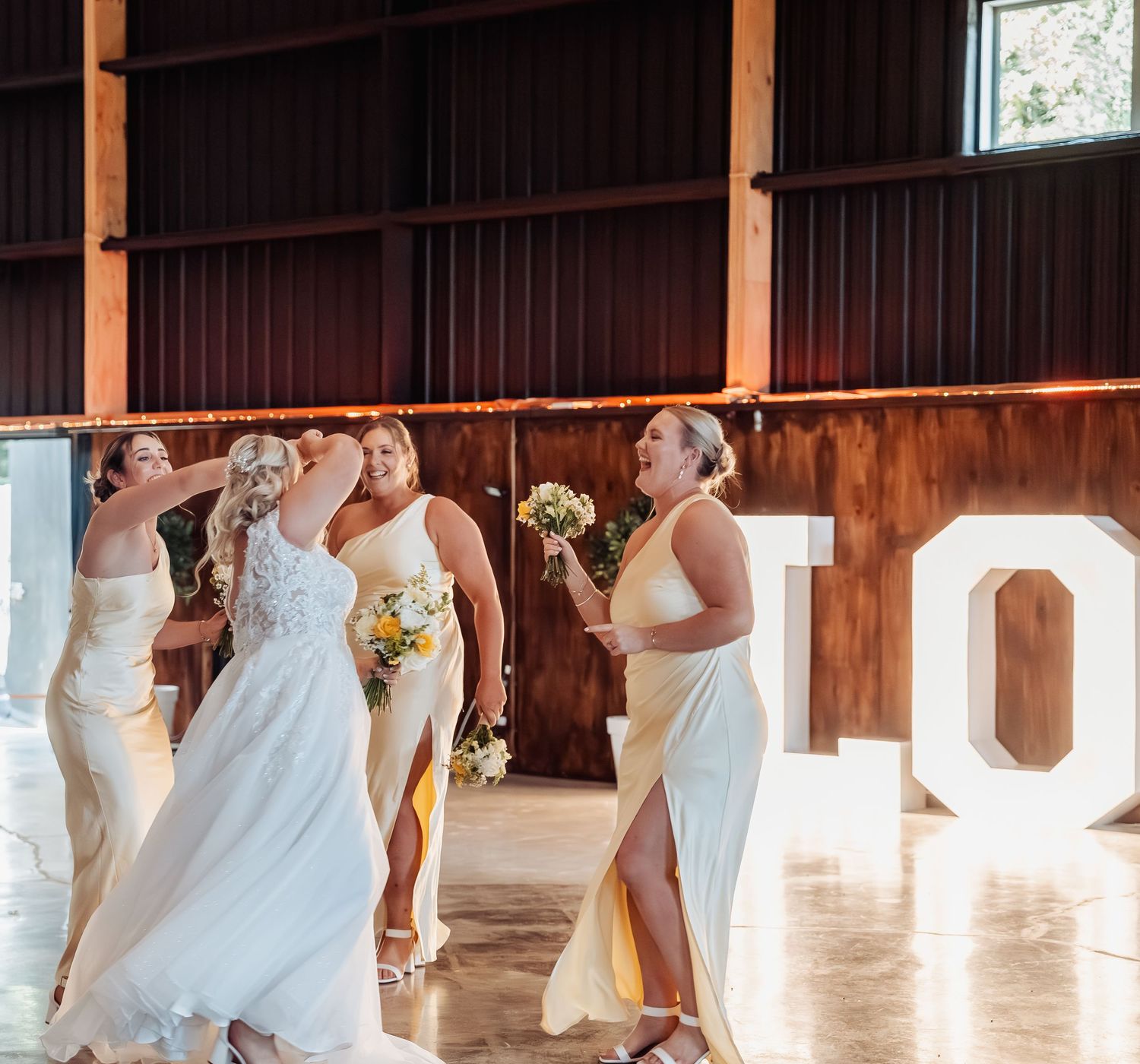 Bride and bridesmaids in yellow dresses holding bouquets inside a rustic barn wedding venue.