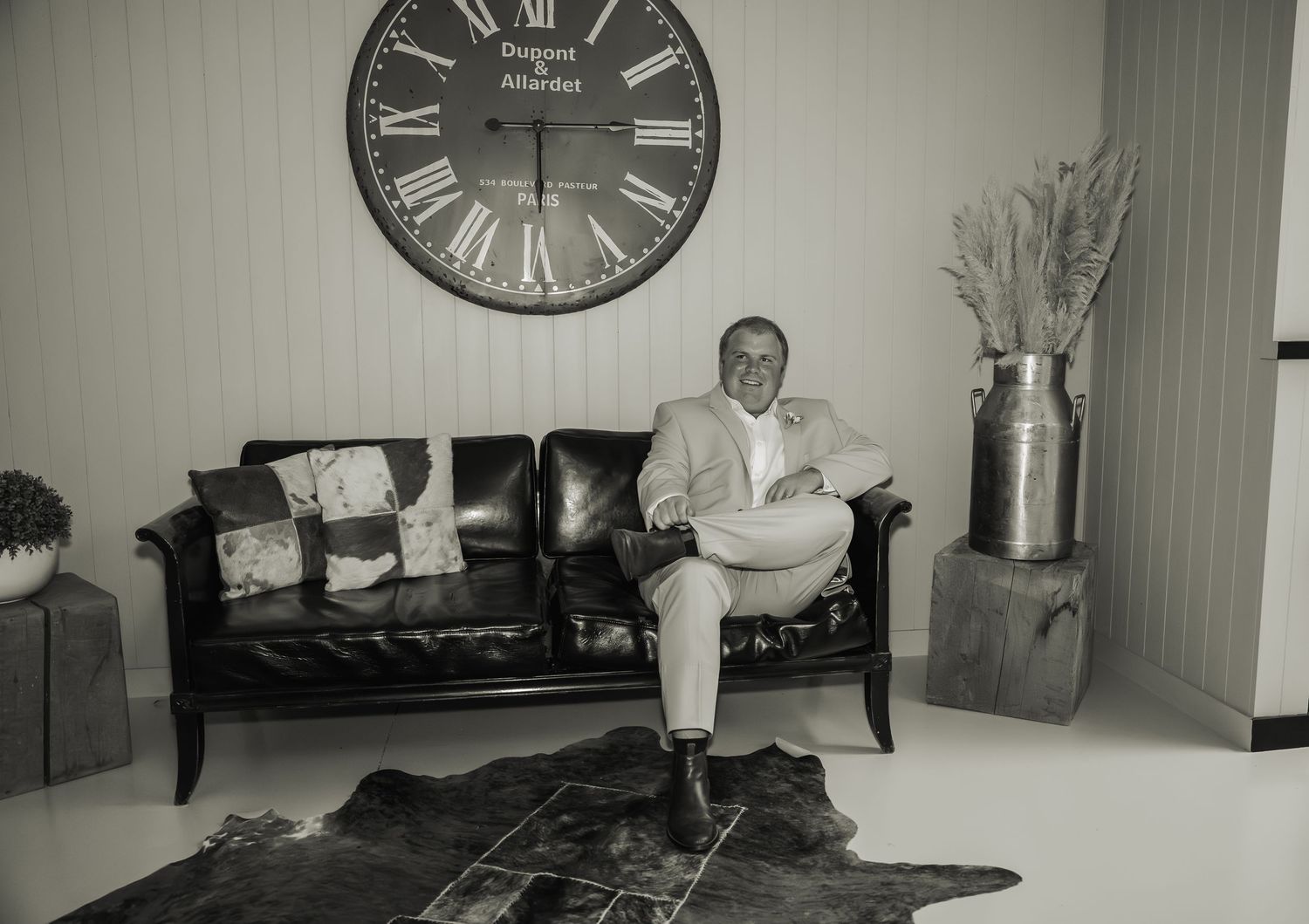 Man in light suit sits on leather sofa beneath large Roman numeral clock in black and white photo.