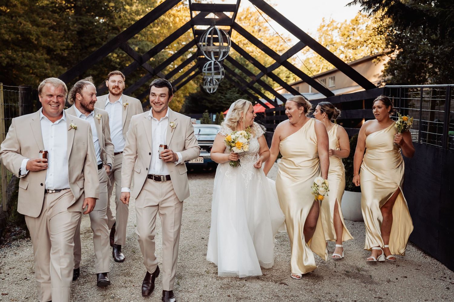 Bride in white gown walks with bridesmaids in gold dresses and groomsmen in tan suits at outdoor wedding.