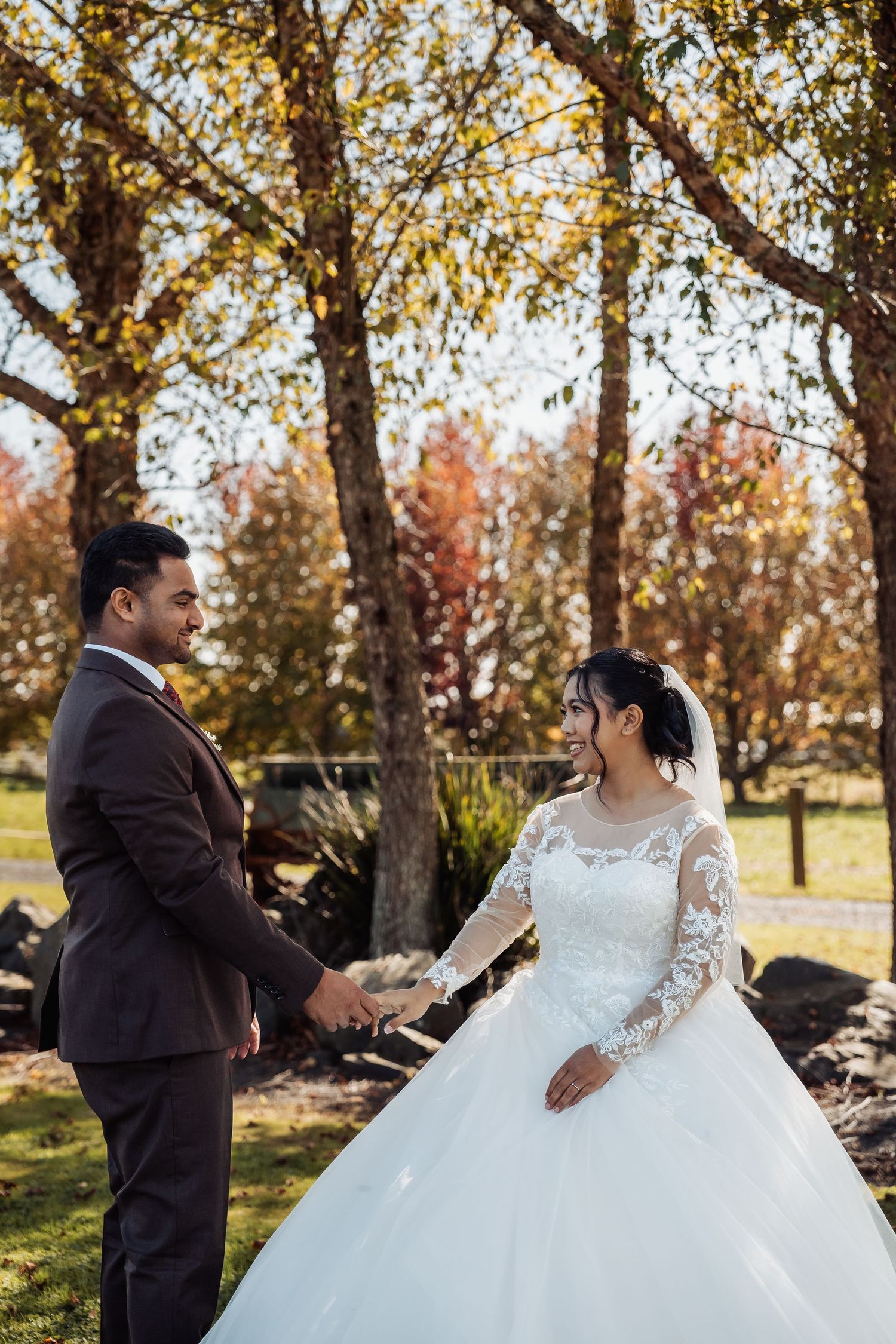 Bride and groom holding hands and smiling at each other in a park with autumn trees in background.