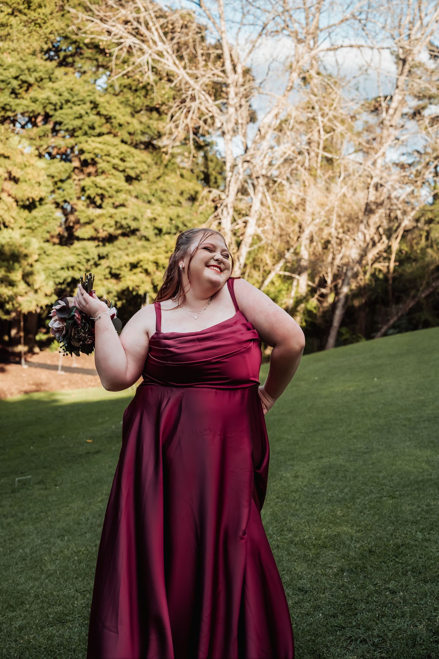 Plus-size bridesmaid in elegant burgundy satin gown poses joyfully on green lawn holding bouquet.