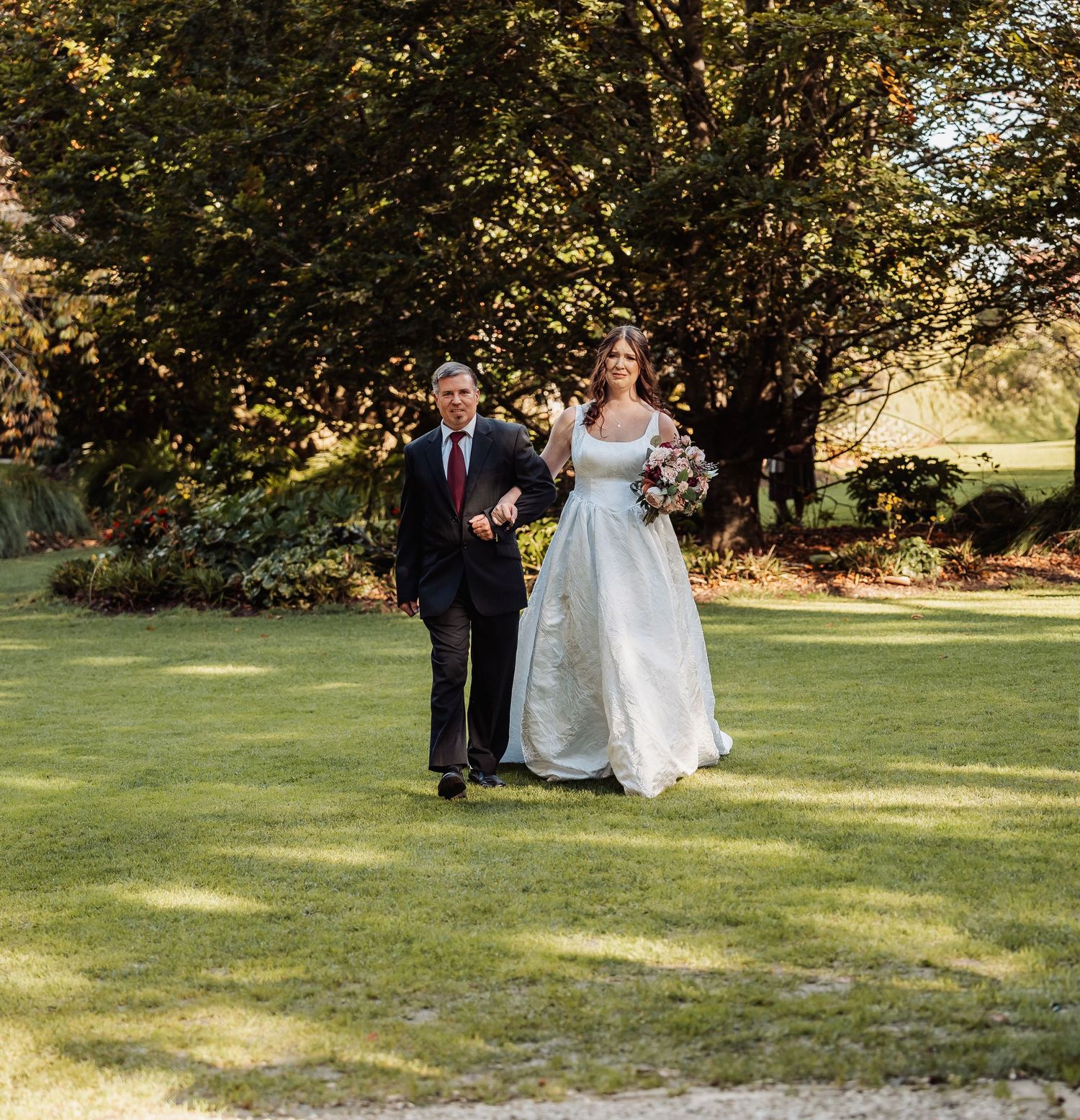 Bride in white gown walks arm in arm with suited gentleman across green lawn at wedding.