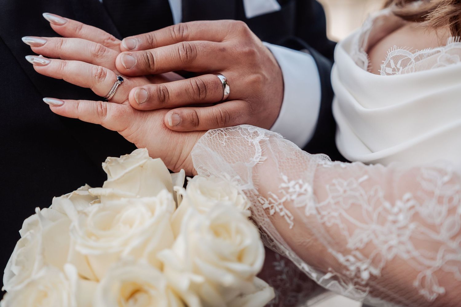 Two hands carefully cut into a three-tiered white wedding cake decorated with delicate floral patterns.