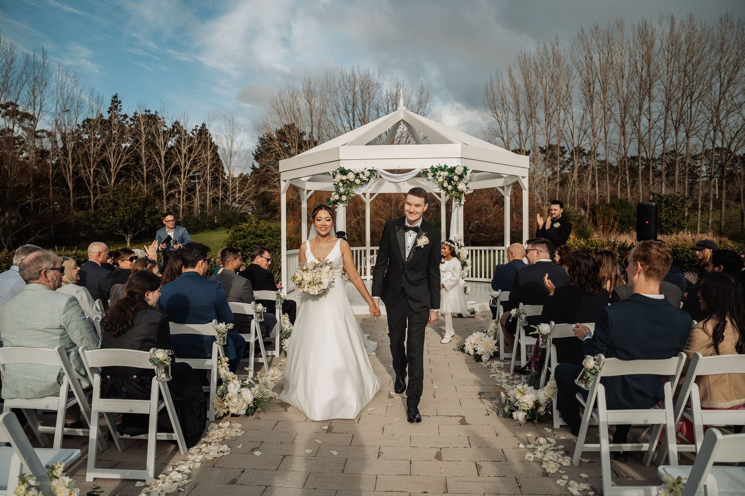 Panoramic view of an outdoor wedding ceremony setup with white chairs and decorated gazebo under cloudy skies.