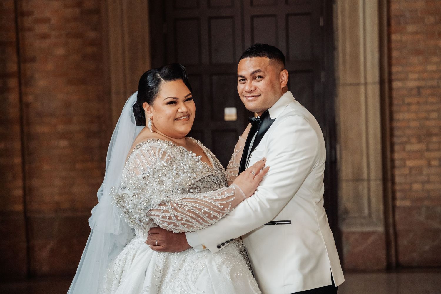 Wedding couple in white attire embracing and smiling in front of a brick building.
