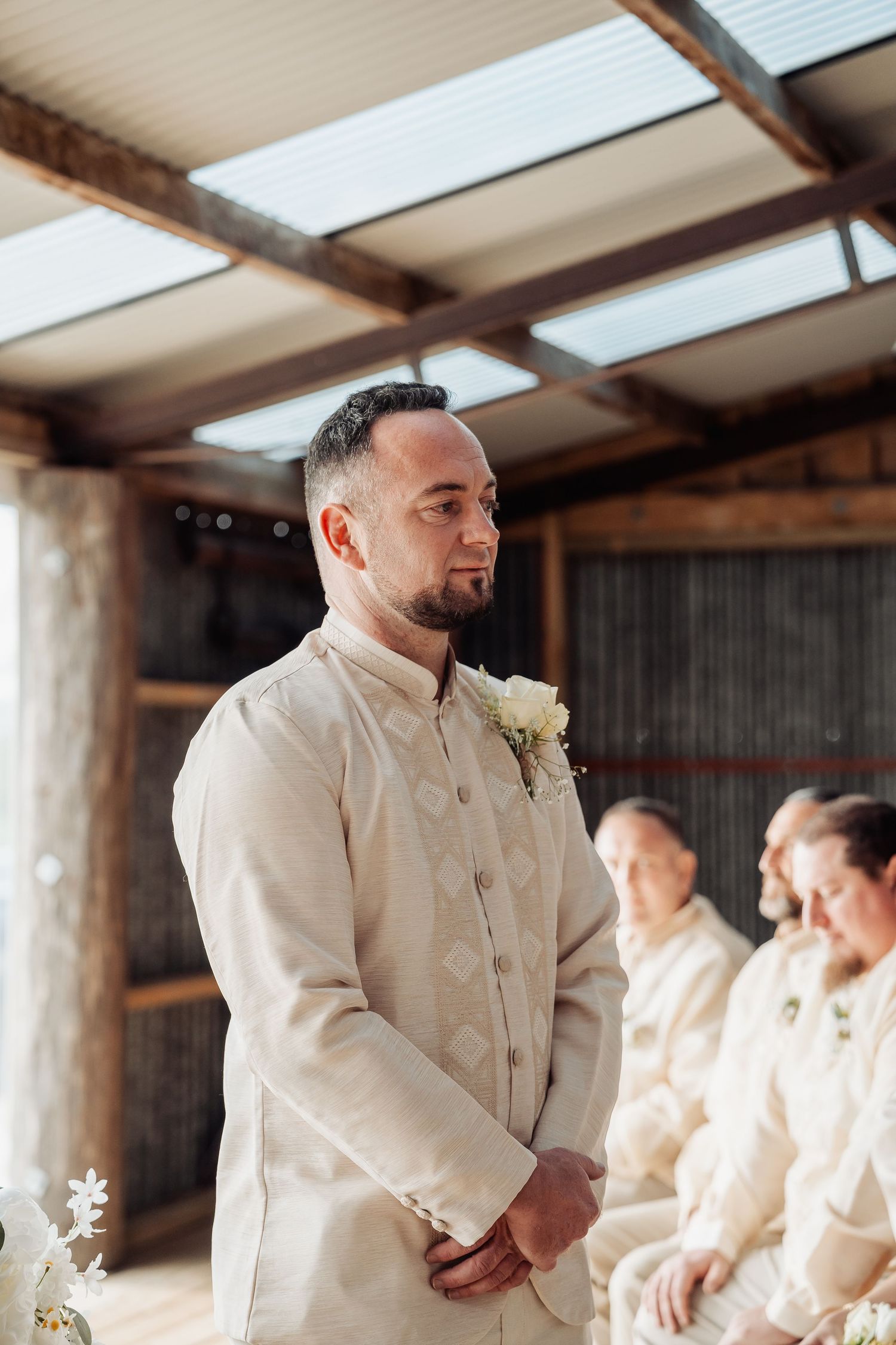 Wedding party members in cream suits stand in a rustic barn venue with wooden beams and skylights.