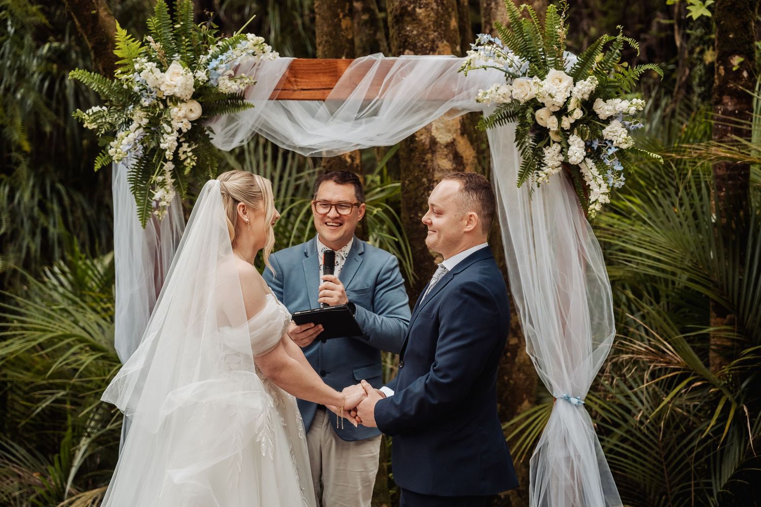 A romantic outdoor wedding ceremony takes place under white drapes surrounded by lush tropical greenery and florals.